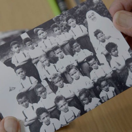 L'image montre une photographie en noir et blanc d'un groupe d'enfants, probablement d'une école ou d'une classe. Les enfants, qui portent des uniformes clairs, sont rassemblés en plusieurs rangées, affichant des expressions diverses. Au centre, une personne vêtue d'une tenue traditionnelle, probablement une enseignante ou une surveillante, se tient parmi eux. L'arrière-plan est flou, mais on peut deviner une ambiance scolaire. Les mains de quelqu'un tiennent la photo avec précaution, indiquant son importance personnelle ou sentimentale.