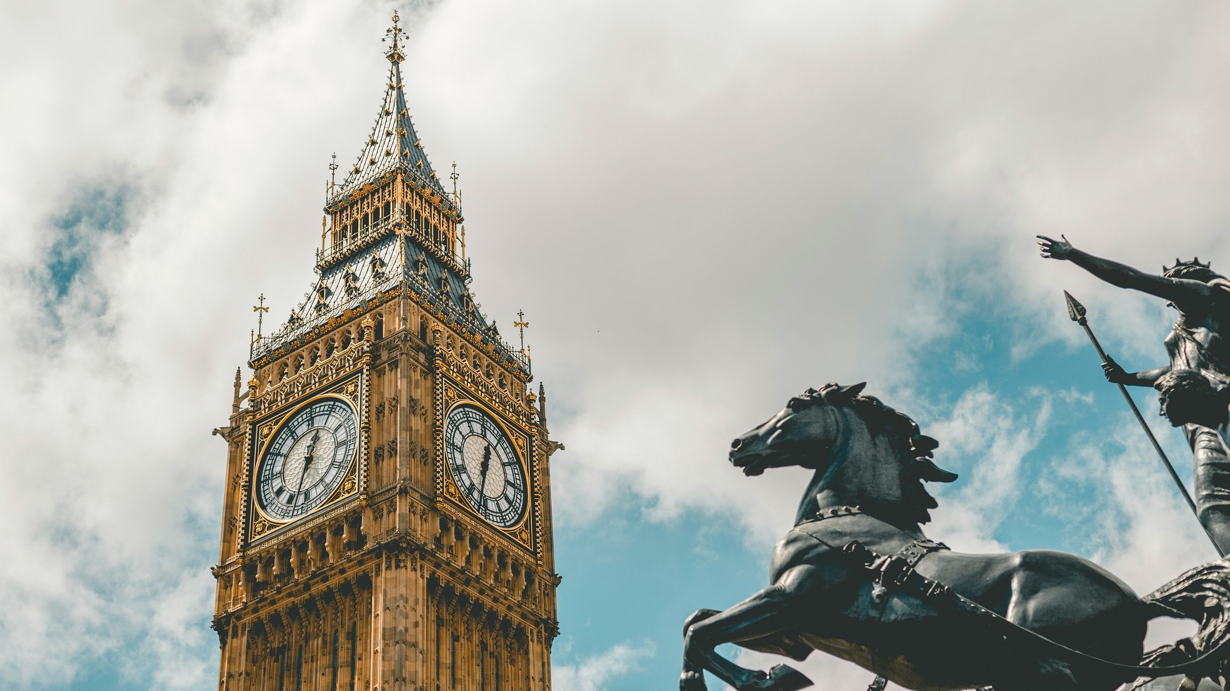 L'image présente une vue majestueuse de la célèbre horloge de Big Ben à Londres. Le bâtiment, en pierre, se distingue par sa hauteur impressionnante et ses ornements dorés. Les deux grands cadrans d'horloge sont visibles, indiquant l'heure avec des chiffres romains. En bas à droite, une statue en bronze d'un cavalier sur un cheval ajoute une touche historique et dynamique à la scène. Le ciel est partiellement nuageux, ce qui crée un contraste intéressant avec l'architecture dorée et les détails du monument. L'ensemble dégage une atmosphère à la fois emblématique et intemporelle.