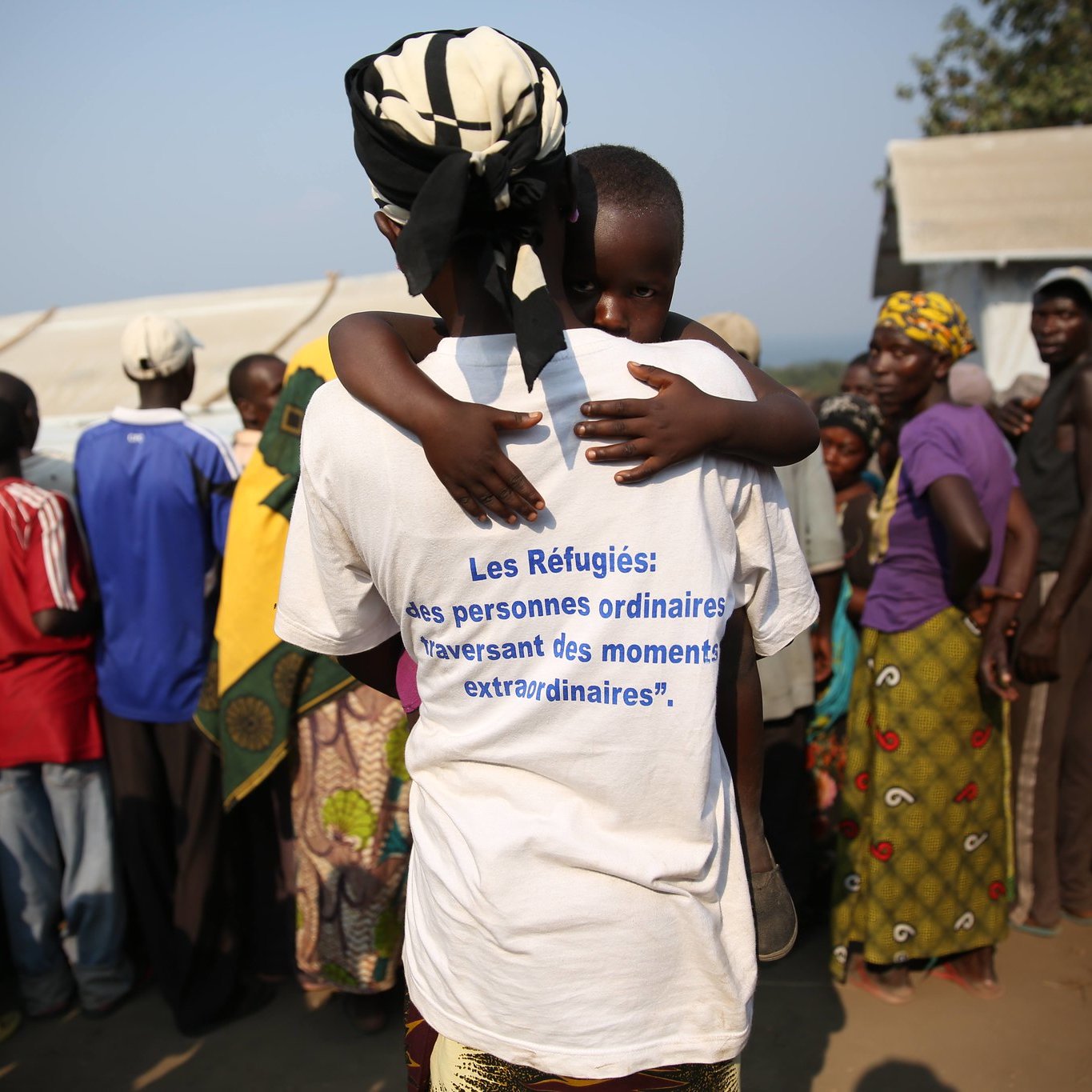 L'image montre un rassemblement de personnes dans un environnement de camp. Au premier plan, une femme, vue de dos, porte un t-shirt blanc portant une inscription en bleu qui dit : « Les Réfugiés : des personnes ordinaires traversant des moments extraordinaires. » Dans ses bras, elle tient un enfant, qui semble se blottir contre elle. En arrière-plan, on peut apercevoir un groupe de personnes, principalement des adultes, ayant des expressions variées. L'ensemble de la scène évoque des thèmes de vulnérabilité et de solidarité. Les vêtements des personnes présentes sont colorés, et l'atmosphère semble à la fois tendue et solidaire. Le décor suggère un campement temporaire, probablement en réponse à une crise.