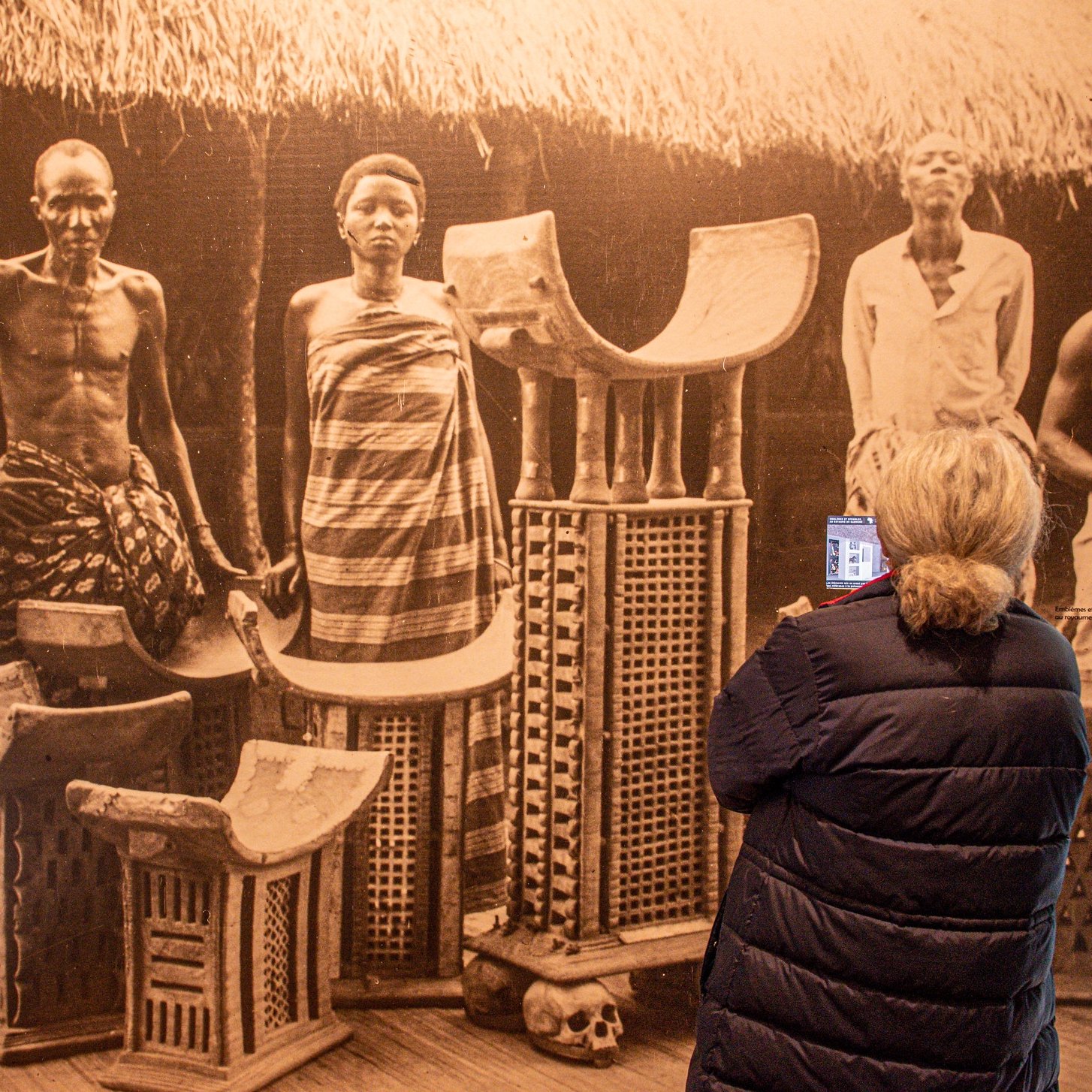 Dans cette image, on aperçoit une scène historique en noir et blanc montrant plusieurs personnes debout, semblant faire partie d'une communauté traditionnelle. Il y a des hommes et des femmes, vêtus de tissus authentiques, avec des poses qui expriment la dignité et la culture. Au centre, on voit des objets artisanaux, comme des chaises ou des tabourets, fabriqués en matériaux naturels, qui portent des motifs décoratifs. À l'arrière-plan, on devine une architecture simple, avec un toit de chaume, créant un cadre authentique. Une femme, de dos, observe cette scène, tenant un appareil dans ses mains, captivée par ce témoignage du passé. L'ambiance générale évoque un sentiment de respect pour les traditions et les histoires des personnes représentées.