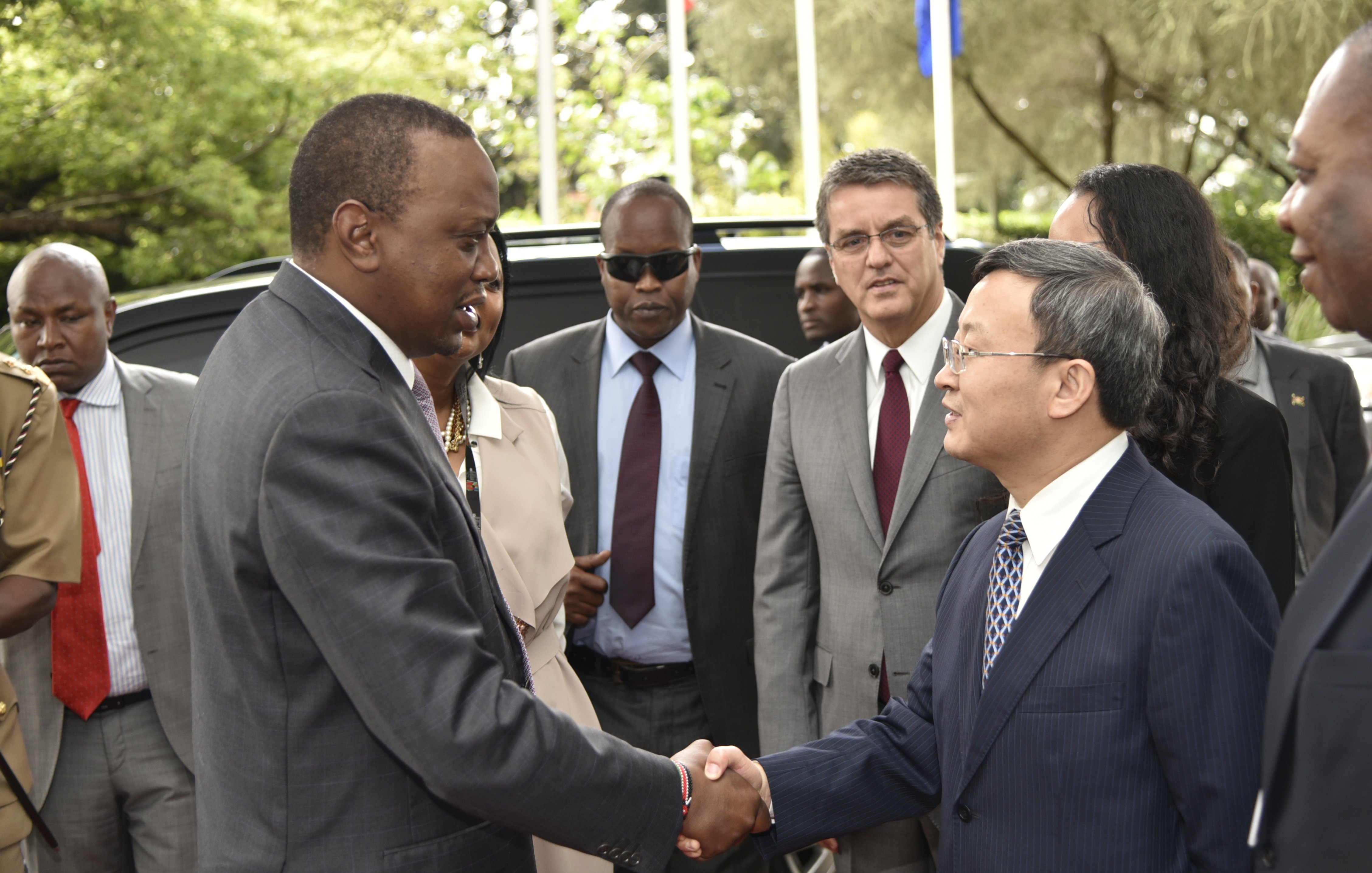 L'image montre une scène de rencontre officielle à l'extérieur, dans un environnement verdoyant avec des arbres et des drapeaux en arrière-plan. Au centre, deux hommes se serrent la main. L'un est habillé en costume sombre, avec une expression sérieuse, tandis que l'autre porte une cravate et un costume clair. En arrière-plan, d'autres personnes observent la rencontre, certaines avec des lunettes de soleil, indiquant un moment formel. L'atmosphère est celle d'une discussion diplomatique.