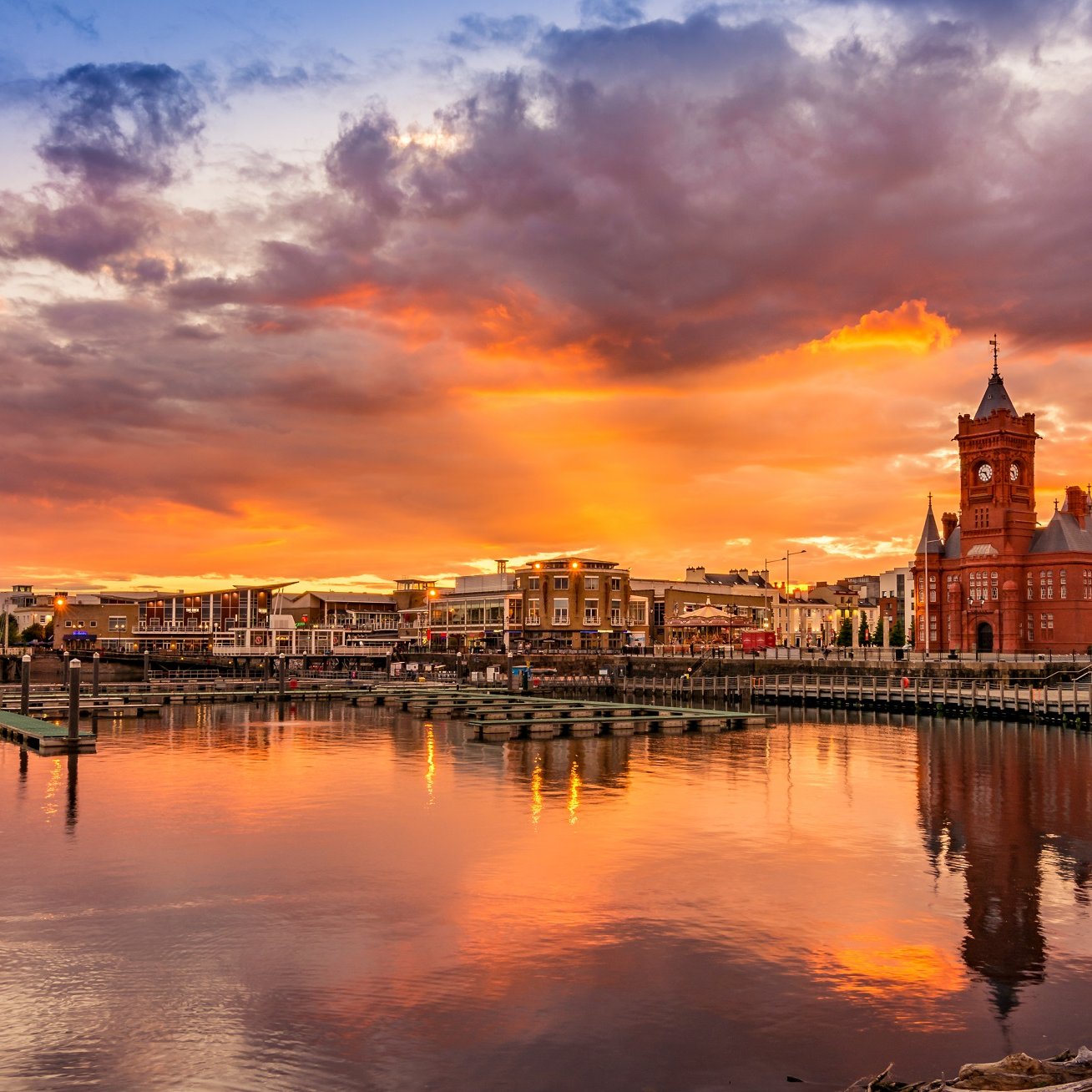 Cette image représente un paysage de bord de mer au coucher du soleil. À l'horizon, un ciel splendide est illuminé par des teintes enflammées de rouge, orange et violet. Le soleil déclinant éclaire les nuages, créant une atmosphère magique. Au premier plan, l'eau calme d'une marina reflète ces couleurs vives, tandis que plusieurs quais et petits bateaux ajoutent à la scène paisible. Sur la rive, un bâtiment architectural distinctif de couleur rouge, avec une tour pointue, attire l'attention, tandis que des arbres et des éléments naturels encadrent le paysage, offrant une ambiance sereine et accueillante.