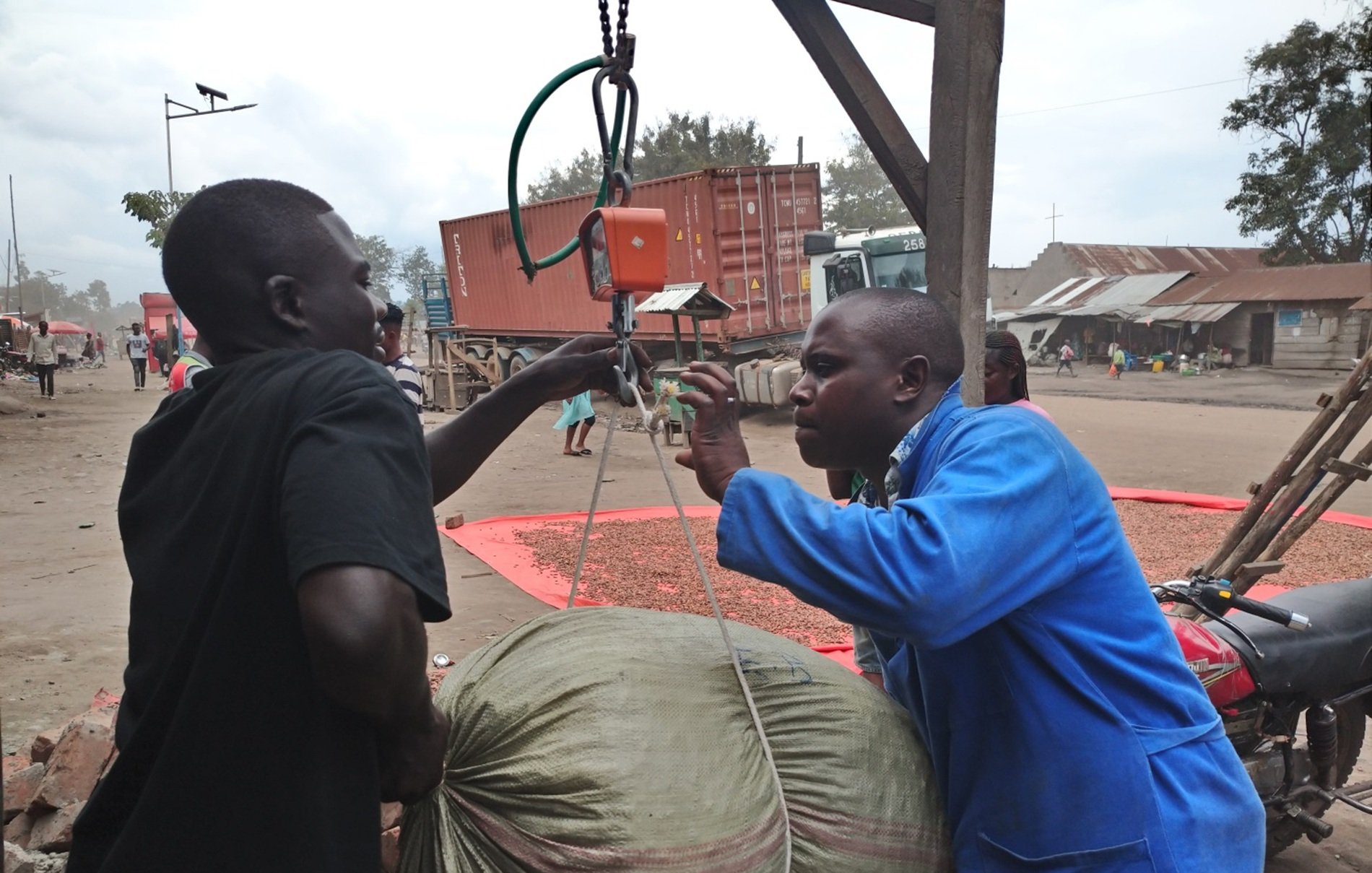 Dans cette image, deux hommes interagissent dans un environnement de marché. L'un des hommes, en tenue bleue, est en train de peser un gros sac à l'aide d'une balance suspendue. Le sac, de couleur beige et légèrement usé, semble lourd. L'autre homme, portant un t-shirt noir, aide à tenir le sac pendant qu'il est pesé. En arrière-plan, on peut apercevoir des conteneurs de transport et des structures typiques d'un marché, avec d'autres personnes qui se déplacent. L'atmosphère est animée, avec des sons de conversation et les bruits d'un marché en pleine activité.