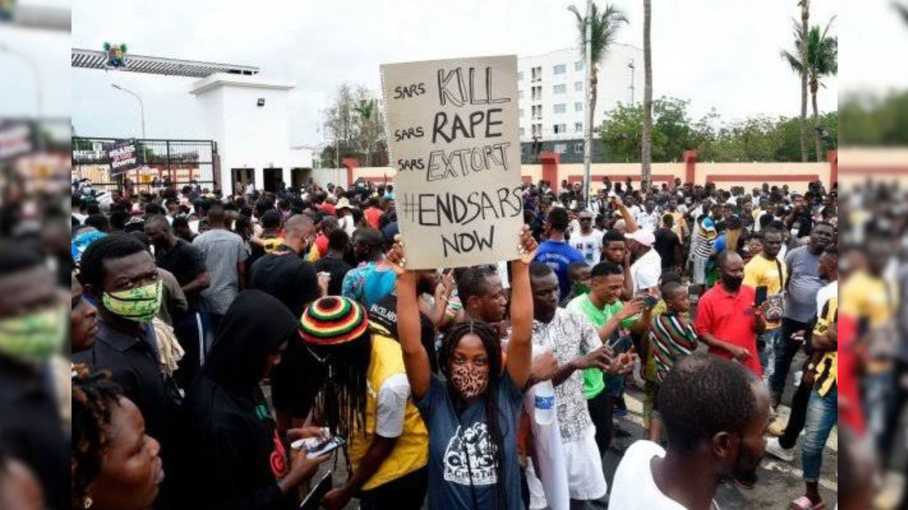 The image depicts a large crowd of people gathered in a protest setting. One individual is holding a sign that reads "KILL RAPE EXTORT END SARS NOW." The atmosphere appears to be serious and unified, with many participants wearing masks. The background includes some trees and buildings, indicating an outdoor urban environment. The protest is likely focused on social issues related to law enforcement.