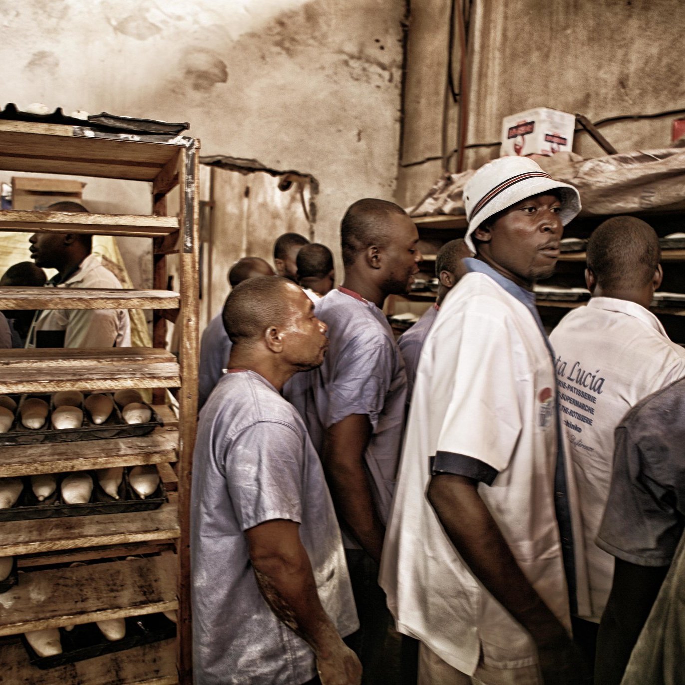 Dans cette image, nous voyons un intérieur sombre et chargé, probablement un espace de travail. Plusieurs hommes se tiennent debout, leurs expressions montrent de l'attention ou de la curiosité. Ils portent des vêtements simples, notamment des chemises et des chapeaux, créant une atmosphère de camaraderie. À l'arrière-plan, on aperçoit des étagères en bois remplie de ce qui semble être des objets enroulés ou des produits. L'éclairage est doux, ajoutant une ambiance de chaleur au lieu. L'ensemble évoque un moment de partage ou de travail collectif.
