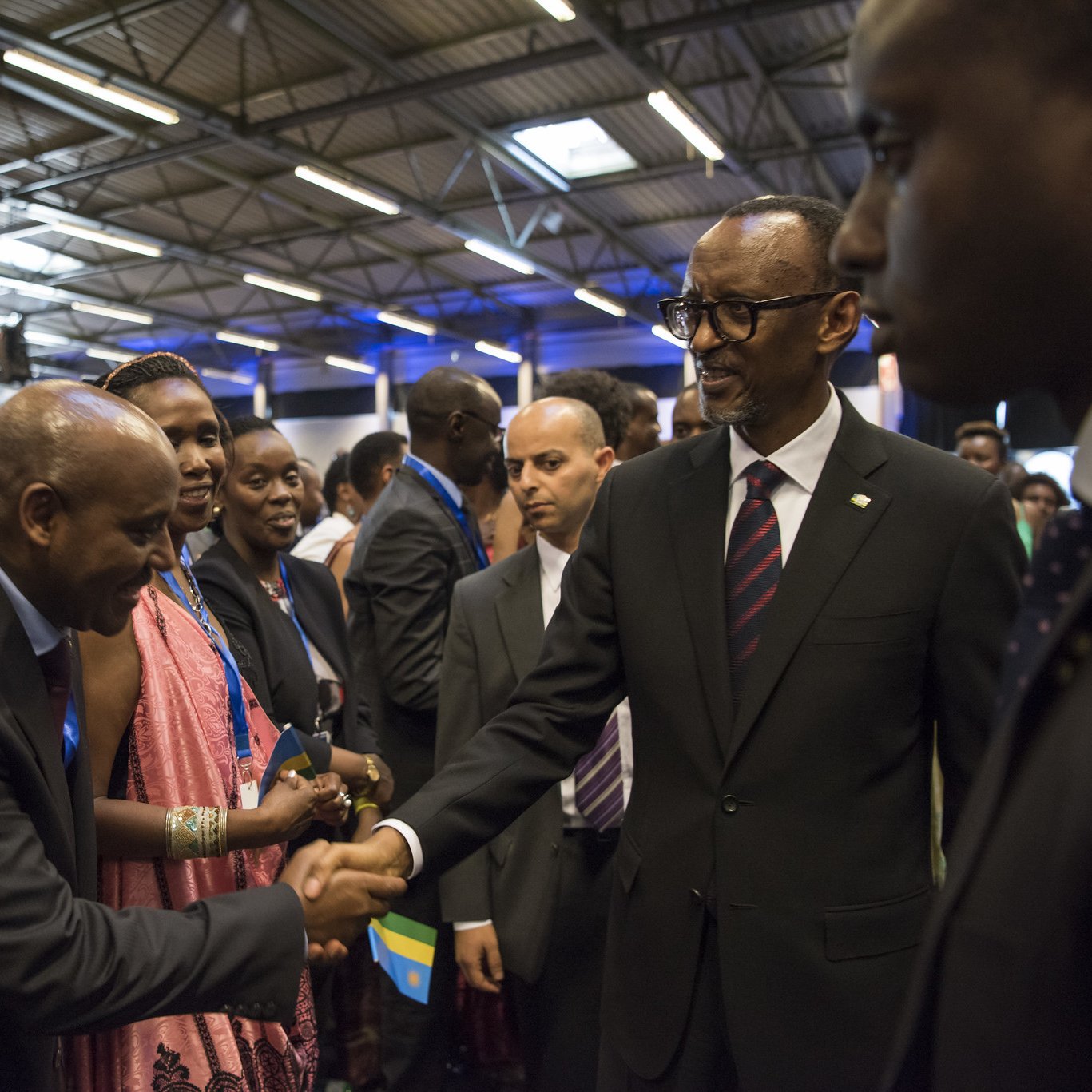 Dans cette image, nous voyons un groupe de personnes rassemblées dans un espace intérieur, probablement lors d'un événement officiel ou d'une conférence. Parmi eux se trouve un homme en costume noir avec une cravate, qui semble être un dirigeant ou une personne importante. Il tend la main pour saluer un autre homme, également en costume. Les autres personnes présentes, principalement habillées formellement, regardent avec intérêt. L'atmosphère est conviviale et respectueuse, avec des sourires et des échanges amicaux. Les murs sont décorés de manière simple, et un éclairage doux illumine la scène.