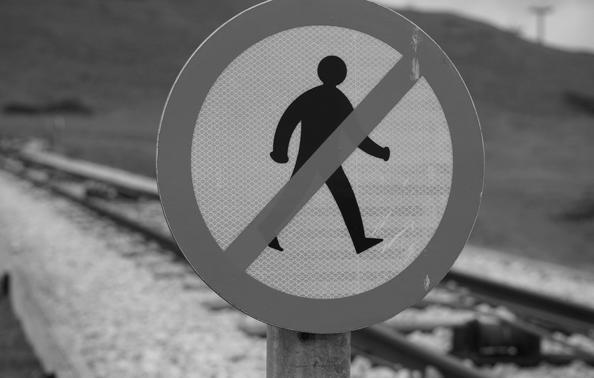 L'image présente un panneau de signalisation circulaire à fond blanc avec une silhouette noire d'une personne marchant. Une barre rouge diagonale traverse le panneau, indiquant qu'il est interdit de marcher ou d'accéder à cette zone. En arrière-plan, on devine des voies ferrées, suggérant que ce panneau est situé à proximité d'une zone ferroviaire, où l'accès est restreint pour des raisons de sécurité. L'image est en noir et blanc, ajoutant un contraste marqué.