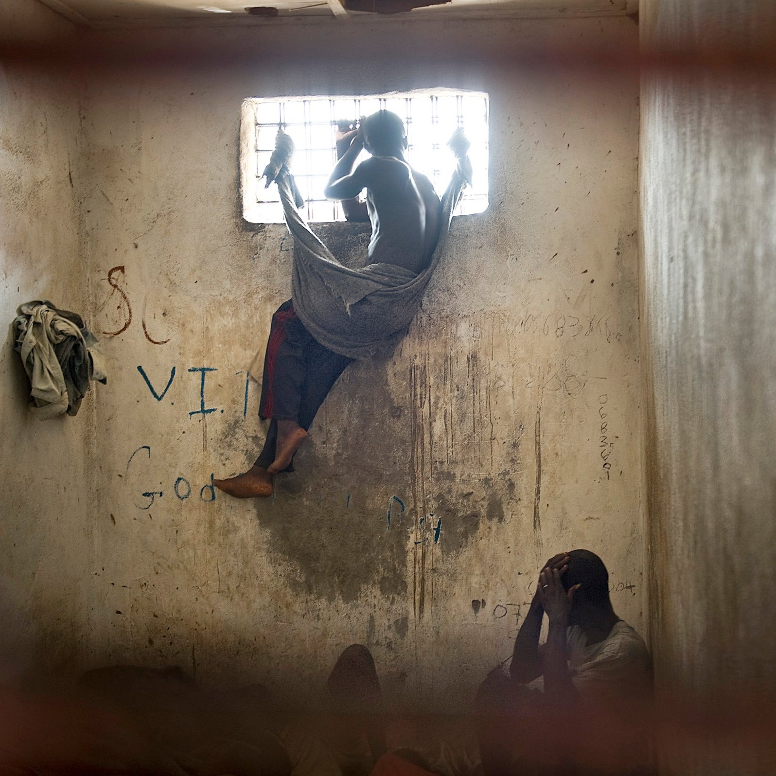 L'image dépeint un environnement sombre et restreint, vraisemblablement une cellule ou une pièce fermée. Au premier plan, deux personnes sont présentes. L'une est assise, visiblement accroupie, la tête entre les mains, adoptant une posture de détresse ou de réflexion. L'autre personne est perchée près d'une fenêtre grillagée, les jambes repliées, enveloppée dans un drap qui semble être une couverture. La lumière qui pénètre par la fenêtre est limitée, créant un contraste entre l'extérieur et l'intérieur sombre de la pièce. Les murs portent des marques, probablement des graffiti, ce qui suggère un environnement négligé. L'atmosphère générale est lourde et empreinte de mélancolie, reflétant un sentiment d'isolement et de désespoir.