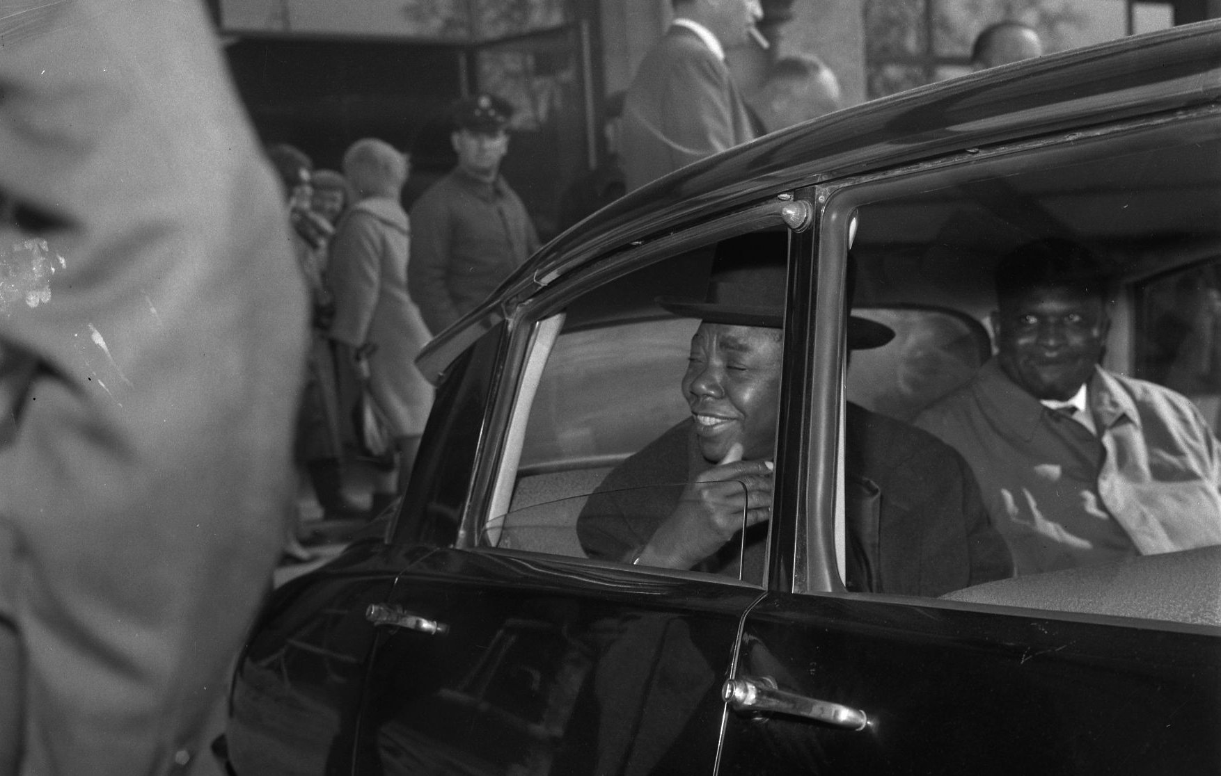 L'image montre l'intérieur d'une voiture noire, où deux hommes sont assis. L'un d'eux, qui se trouve à l'avant, semble souriant et engageant, habillé d'un costume élégant et portant un chapeau. Son expression indique qu'il est en train de parler ou de rire. À l'arrière de la voiture, l'autre homme a une expression plus neutre, vêtu également d'un manteau. À l'extérieur de la voiture, on peut apercevoir une foule de personnes en arrière-plan, certaines s'attardant dans un hall ou une gare d'arrivée. L'atmosphère semble animée, suggérant un moment de retrouvailles ou d'attente.