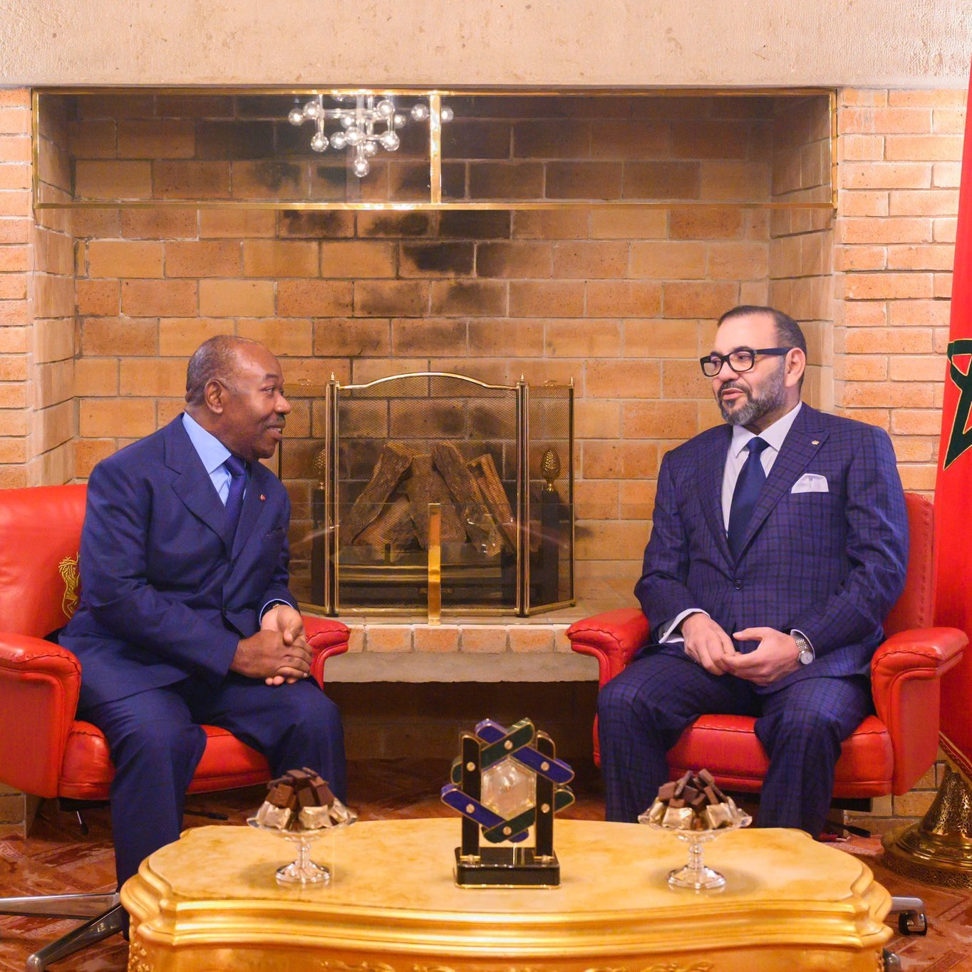 L'image montre deux hommes assis face à face dans un salon chaleureux et élégant. À gauche, un homme en costume bleu, avec une cravate, est légèrement incliné vers l'avant, écoutant attentivement. À droite, un autre homme porte un costume à carreaux bleu foncé et une cravate assortie, affichant une expression sérieuse tout en participant à la conversation. Derrière eux, on peut voir un mur en briques claires et une cheminée décorative. Il y a également des éléments de décoration comme des fauteuils rouges et un tableau avec un drapeau marocain sur le côté droit. Une lumière douce illumine la pièce, créant une atmosphère conviviale.