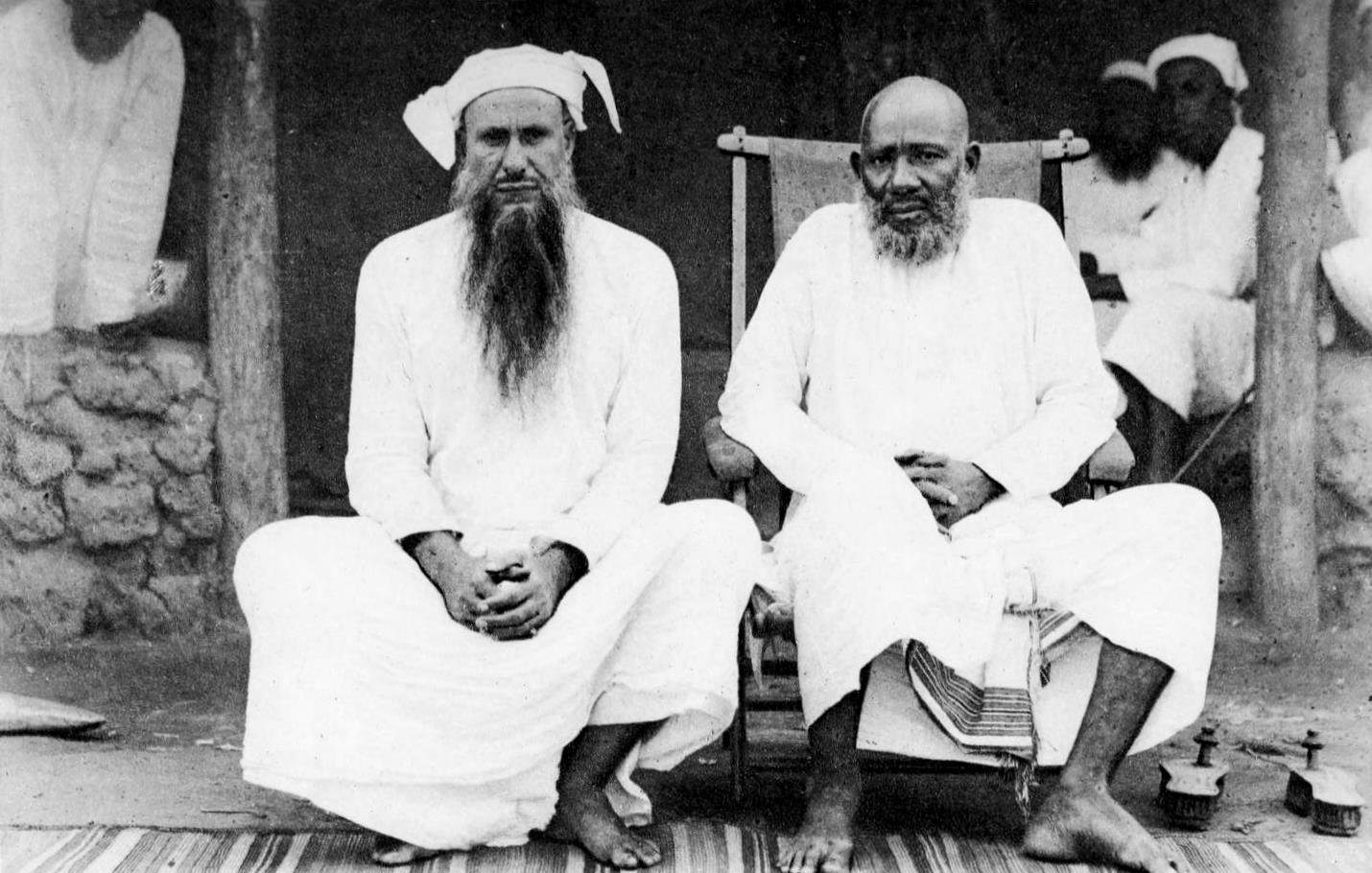 L'image montre deux hommes assis côte à côte sur un tapis au sol. L'homme de gauche, portant une longue barbe blanche et un turban, est habillé d'une tunique blanche. À sa droite se trouve un autre homme, également en tunique blanche, qui semble plus âgé, avec une barbe grise. Ils affichent tous deux une expression calme et sereine. En arrière-plan, on peut distinguer des silhouettes d'autres personnes floues et un environnement rural. Ces détails donnent une impression de tranquillité et de tradition.