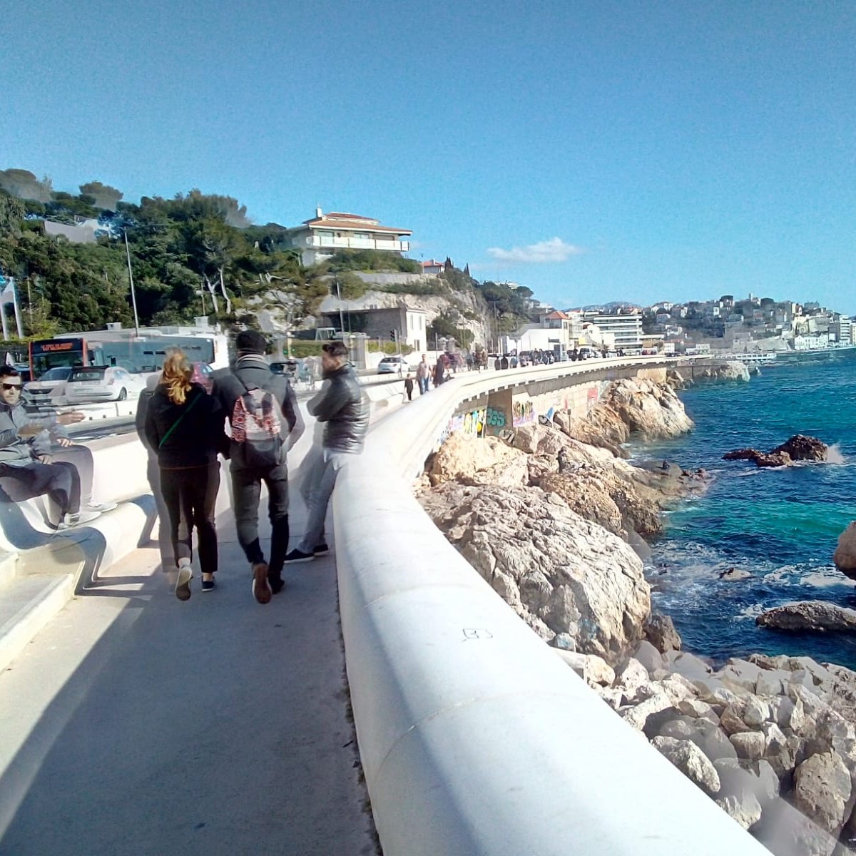 Imaginez-vous sur une promenade longeant la mer. Sur votre droite, vous pouvez entendre le bruit des vagues qui s'écrasent doucement contre les rochers, tandis que l'odeur salée de l'océan flotte dans l'air. Le vent léger caresse votre visage et apporte une sensation de fraîcheur. À votre gauche, des bâtiments se dressent, certains sont des maisons et d'autres des commerces. Vous pouvez sentir l'animation de la ville avec des groupes de personnes qui marchent, certains discutant joyeusement. La promenade est large et bien aménagée, invitant à la flânerie. Au loin, le paysage est ponctué d'arbres et de collines, avec quelques nuages blancs dégradant le bleu du ciel. La lumière du soleil illumine tout le paysage, créant une ambiance chaleureuse. L'ensemble évoque une belle journée au bord de la mer, propice à la détente et au plaisir des sens.