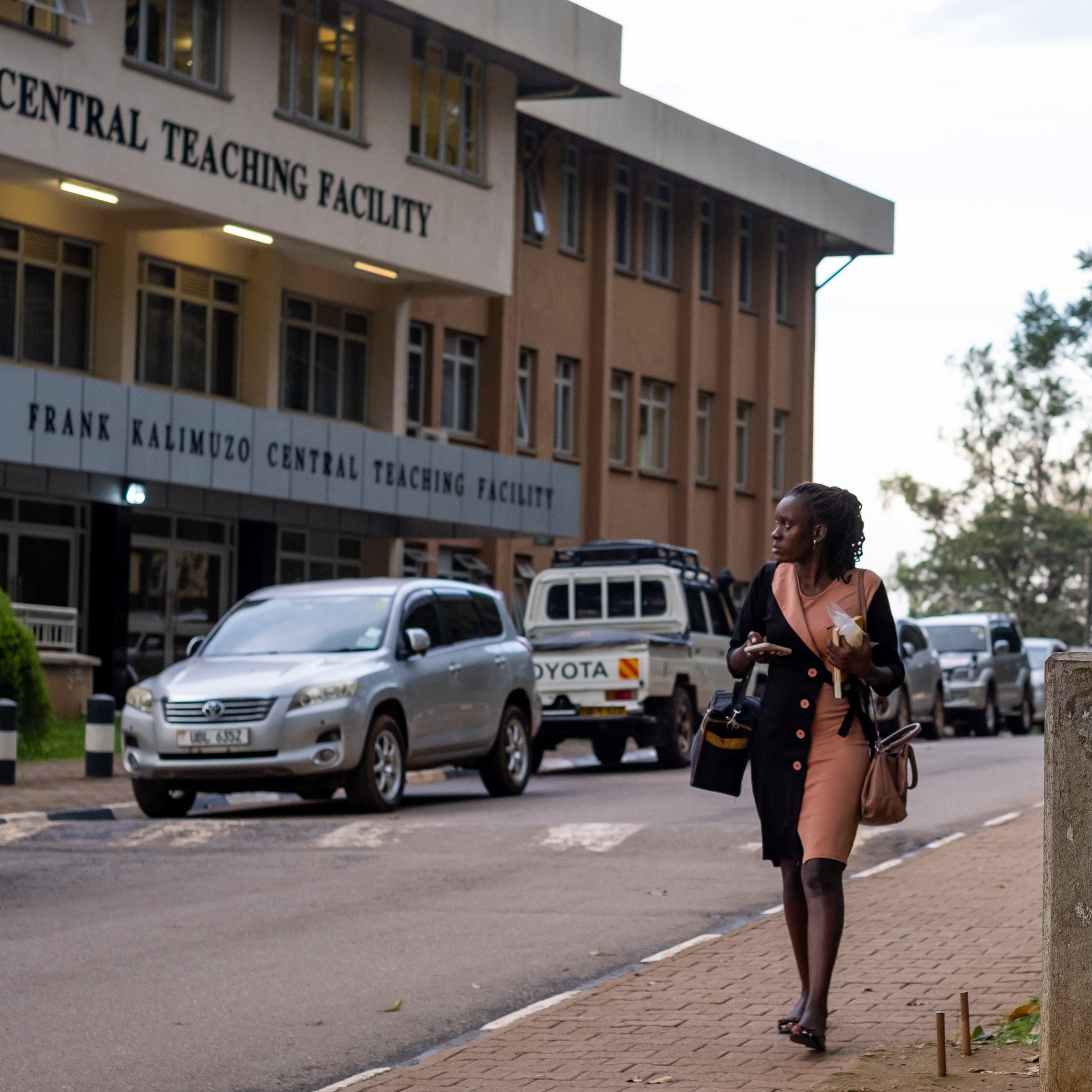 Dans cette image, on voit une scène urbaine près d'un bâtiment nommé "Frank Kalimuzo Central Teaching Facility". La structure est moderne avec plusieurs fenêtres et un design clair. Sur le trottoir, une femme vêtue d'une robe rose marche, portant une sacoche. À l'arrière-plan, on aperçoit quelques véhicules, dont une voiture blanche, et des arbres qui apportent une touche de verdure à l'environnement. Des panneaux sur un mur en béton à droite attirent l'attention avec des avertissements écrits en grandes lettres. L'ambiance est calme, suggérant un moment de transition entre la journée et le soir.