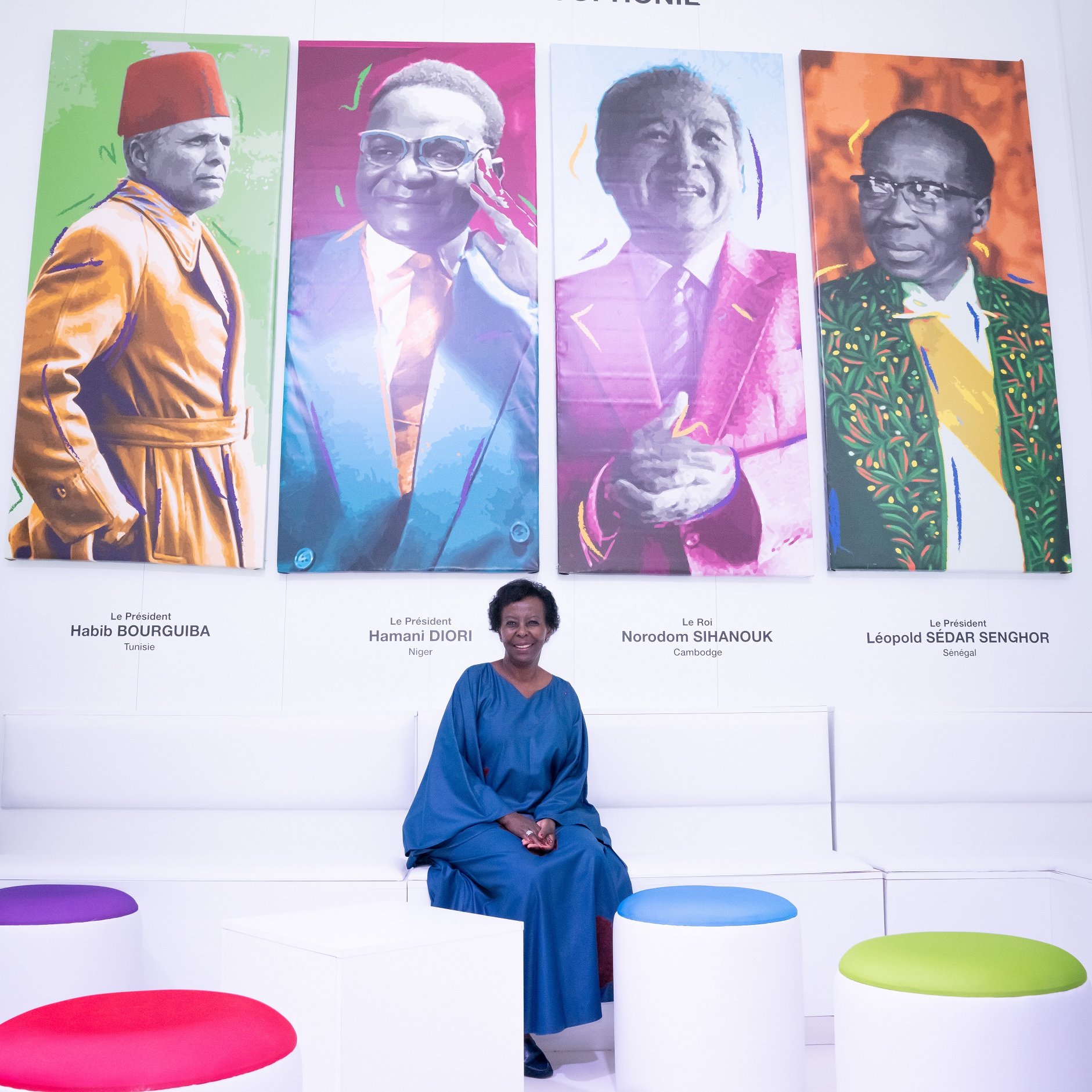 L'image présente une femme assise sur un banc dans une galerie moderne. Derrière elle, il y a quatre portraits colorés de figures politiques africaines. Chaque portrait est visiblement distinct, avec des couleurs vives, représentant des hommes portant des costumes traditionnels ou modernes. La femme, vêtue d'une robe bleu foncé, sourit légèrement et semble apprécier l'exposition. Le décor est minimaliste, avec des sièges blancs et des poufs de différentes couleurs qui ajoutent une touche de dynamisme à l'ensemble. L'ambiance générale est accueillante et artistique, invitant à la réflexion sur l'histoire politique de l'Afrique.