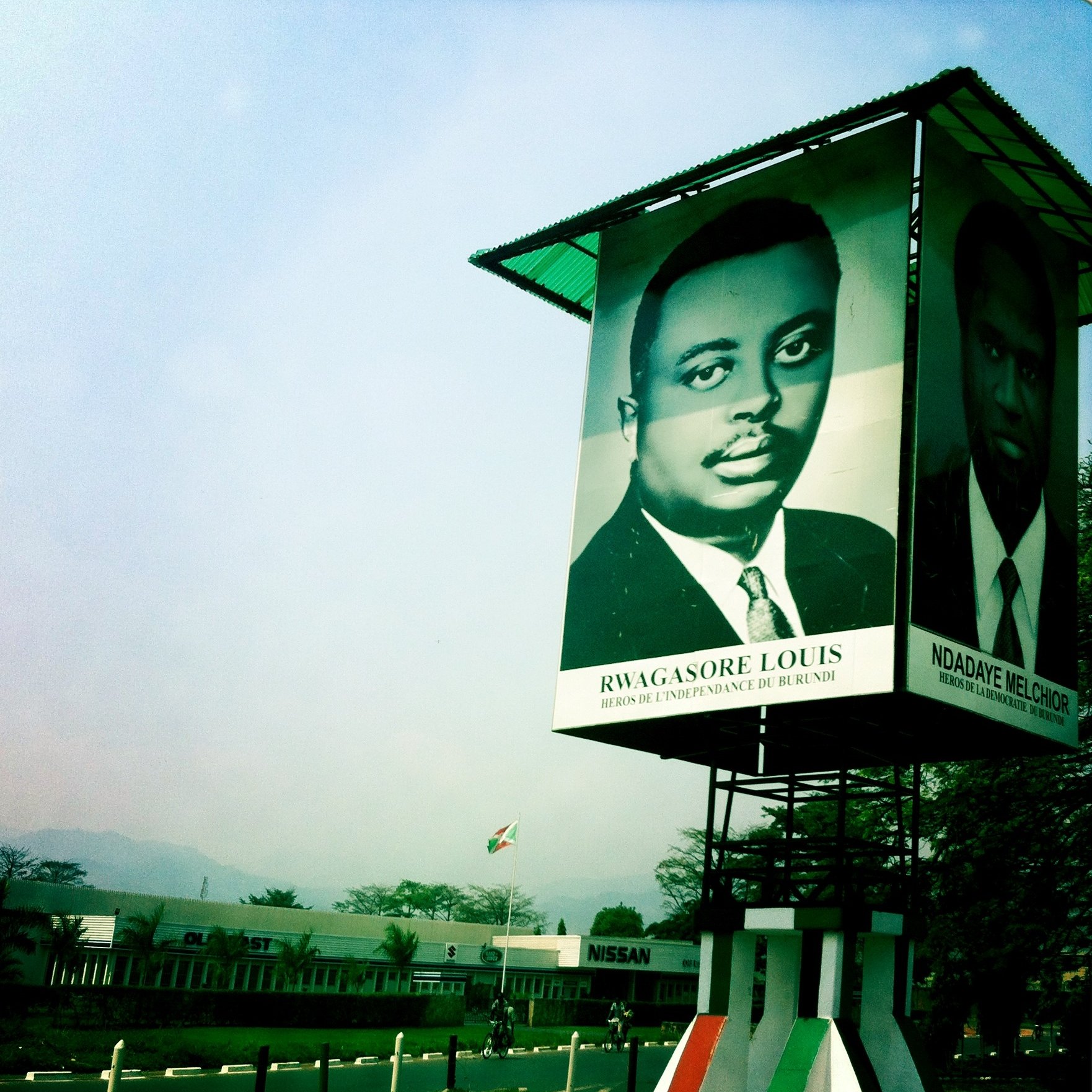 Cette image montre un grand panneau publicitaire ou une affiche située dans un environnement urbain. Sur le panneau, il y a un portrait en noir et blanc d'un homme, sous lequel est inscrit son nom : "RWAGASORE LOUIS", avec la mention "Héros de l'indépendance du Burundi". À côté de ce portrait, on aperçoit un autre visage (sans nom visible). Le fond de l'image présente un ciel légèrement brumeux et des bâtiments en arrière-plan, ce qui indique un paysage urbain. L'ambiance de l'image est à la fois historique et commémorative, mettant en avant des figures importantes pour le Burundi.