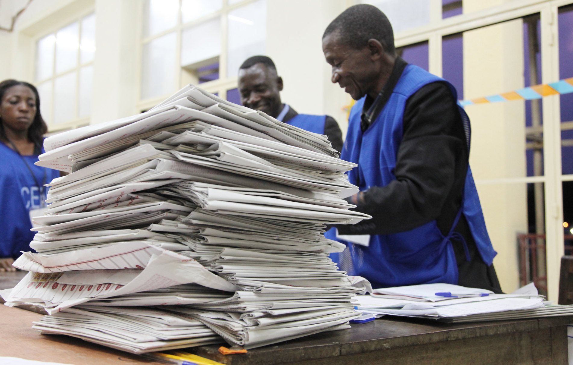 L'image montre une scène à l'intérieur d'un bureau de vote. Au premier plan, on aperçoit une grande pile de documents, probablement des bulletins de vote ou des formulaires électoraux, soigneusement empilés sur une table en bois. À l'arrière-plan, trois personnes en gilets bleus se trouvent près de la table, semblant discuter ou examiner les papiers. L'atmosphère est concentrée, suggérant une activité importante liée à un scrutin. La lumière éclaire la pièce, créant une ambiance sérieuse et professionnelle.