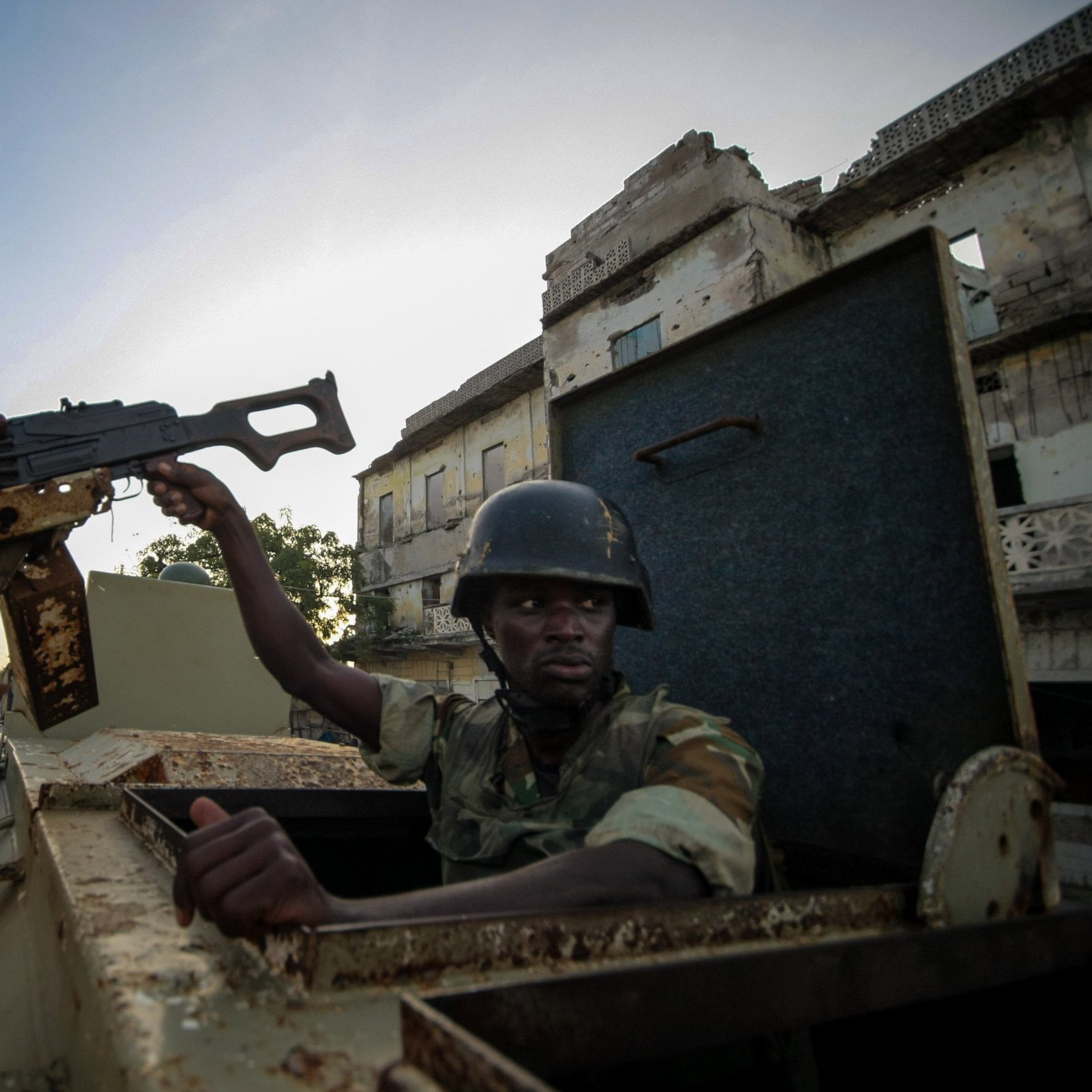 L'image montre un soldat assis dans un véhicule militaire. Il est vêtu d'un uniforme camouflage et porte un casque. Le soldat tient un fusil d'assaut, que l'on peut imaginer prêt à être utilisé. En arrière-plan, on aperçoit des bâtiments en ruine, suggérant que l'environnement est marqué par des signes de conflit. La lumière est douce, indiquant peut-être un lever ou un coucher de soleil. L'atmosphère dégage un sentiment de tension et de vigilance dans un cadre urbain dégradé.