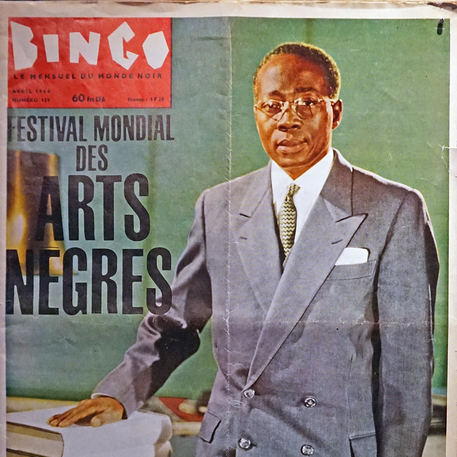 L'image représente une couverture de magazine intitulée "Bingo", datée d'avril 1966. Au premier plan, un homme debout est habillé d'un costume clair, avec une chemise blanche et une cravate sombre. Il porte des lunettes et affiche une expression sérieuse. L'arrière-plan est flou, mais semble être un environnement sobre, mettant en valeur le sujet. En haut de la couverture, on peut lire "FESTIVAL MONDIAL DES ARTS NÈGRES", ce qui évoque un événement culturel important. Les couleurs du magazine sont douces, ajoutant une touche d'élégance à l'ensemble.