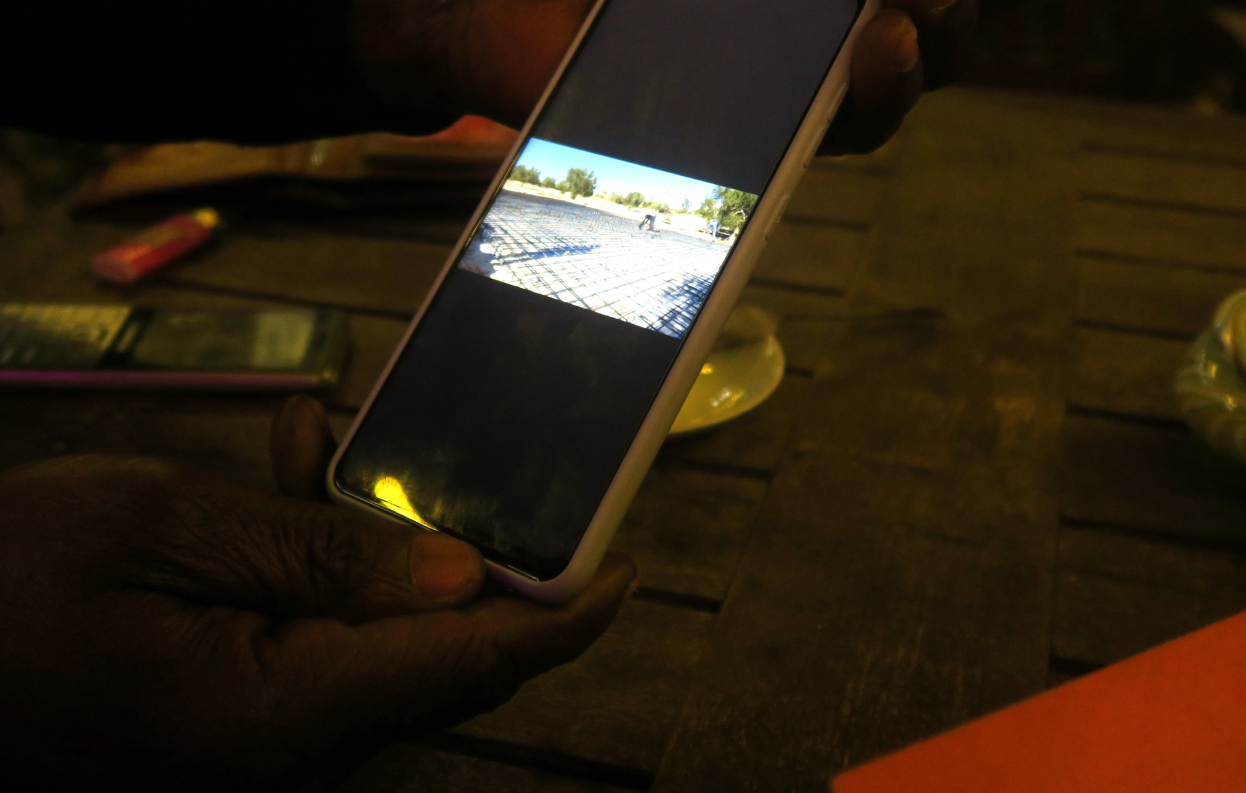 L'image montre une main tenant un téléphone portable, qui affiche une photographiée (ou une vidéo) d'un paysage. À l'arrière-plan, on peut imaginer une scène en extérieur, probablement un terrain ou une route. La lumière dans la pièce est tamisée, et il y a une table en bois visible. Sur la table, on peut apercevoir quelques objets, comme un petit paquet de cigarettes et un plateau. L'ambiance semble tranquille et décontractée, suggérant un moment de détente ou de partage.