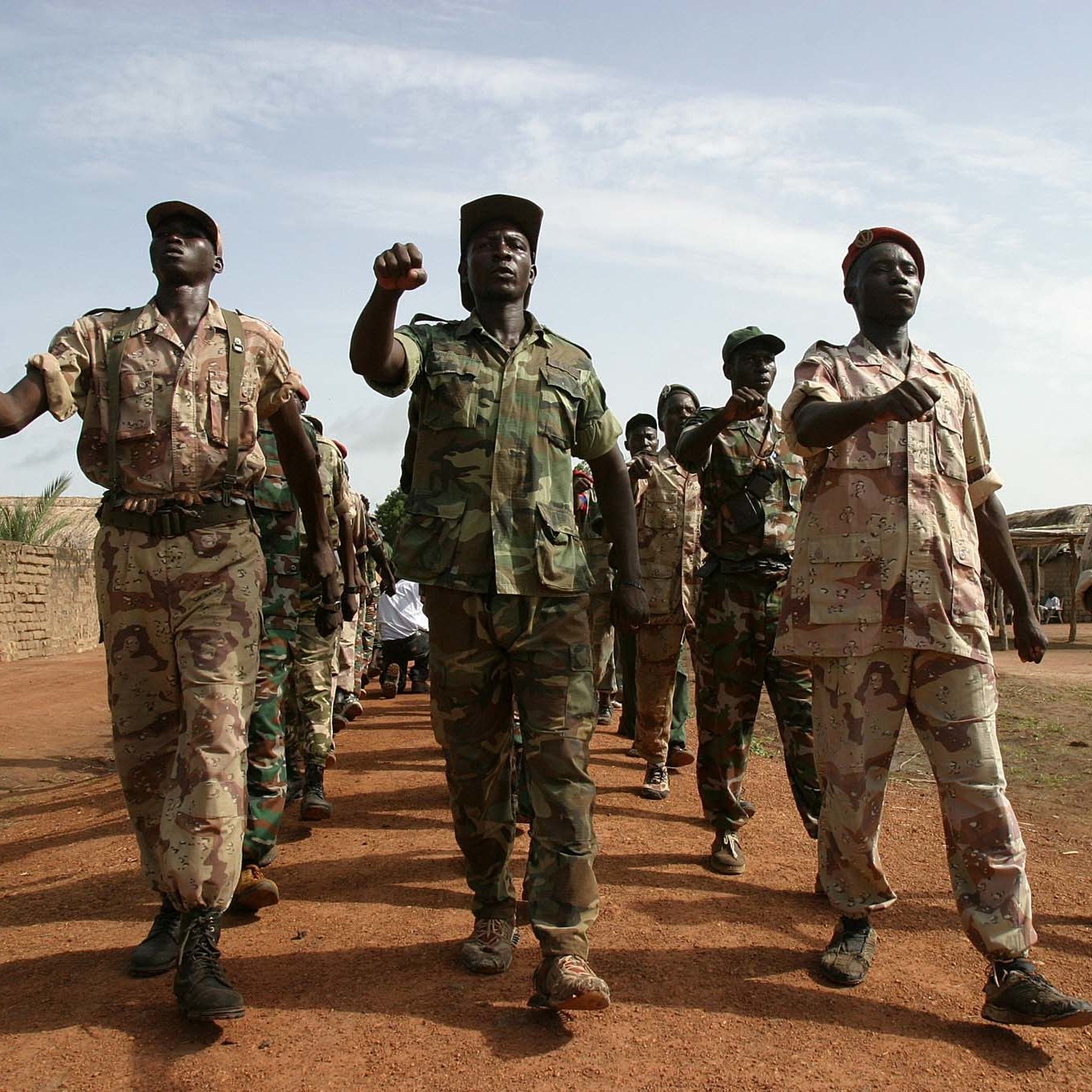 L'image montre un groupe de soldats marchant en formation sur un sol poussiéreux. Ils portent des uniformes militaires variés, allant du camouflé au beige, qui suggèrent une ambiance militaire. Certains soldats brandissent le poing en signe de détermination, tandis que d'autres avancent d'un pas ferme. En arrière-plan, on peut voir des tentes militaires ou des structures rudimentaires, suggérant un camp. Le ciel est légèrement nuageux, laissant passer quelques rayons de soleil. L'atmosphère générale évoque la discipline et la camaraderie au sein de ce groupe.