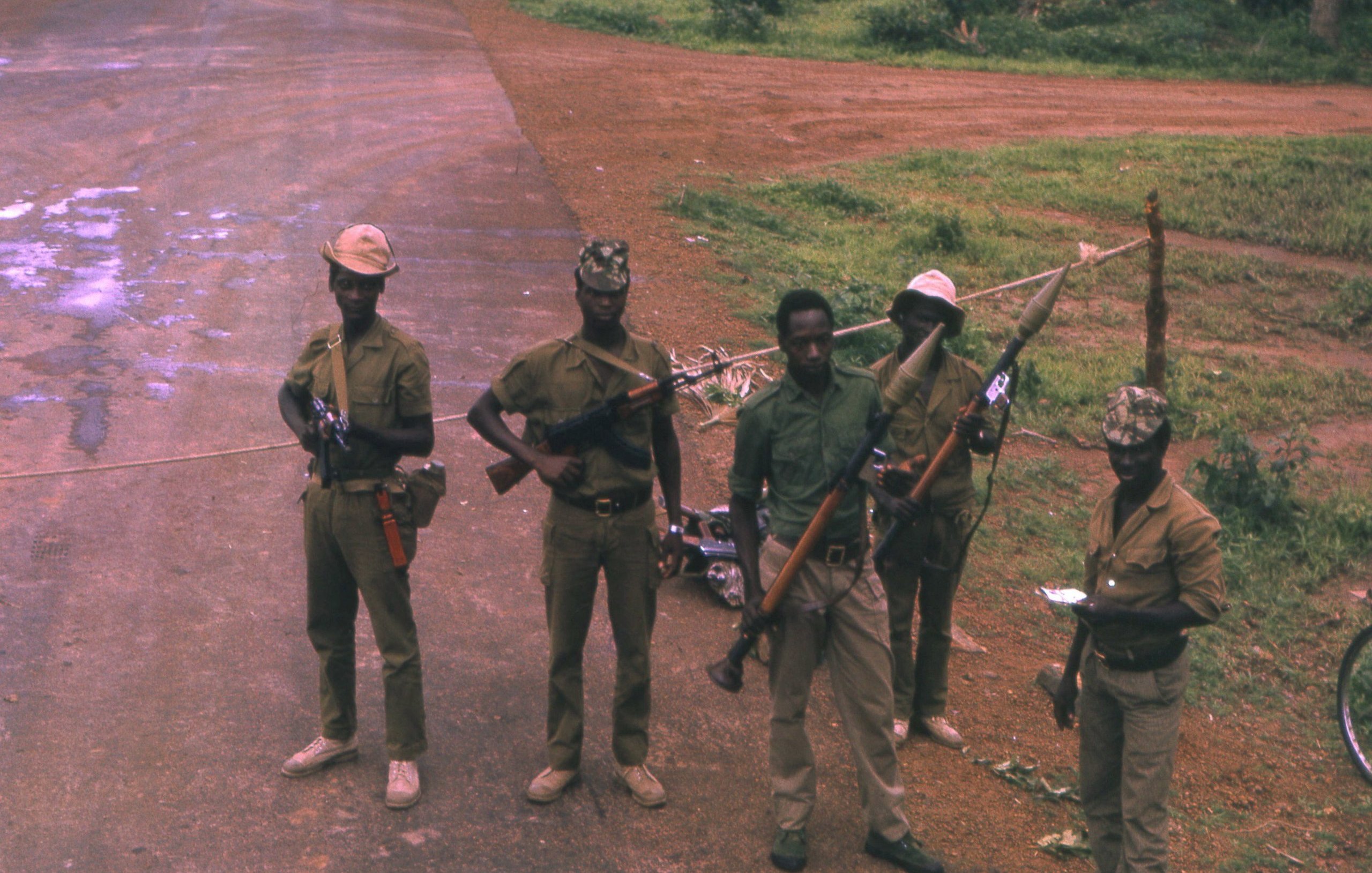 L'image montre un groupe de cinq soldats en uniforme, debout sur une route non pavée. Ils portent des chapeaux, et chacun d'eux tient une arme à feu. Le sol est poussiéreux, avec quelques zones humides visibles. En arrière-plan, on voit une végétation luxuriante, typique d'un environnement tropical. Les soldats affichent des postures sérieuses, et certains d'entre eux paraissent concentrés sur leur environnement. Cette scène suggère un contexte de sécurité ou de patrouille.