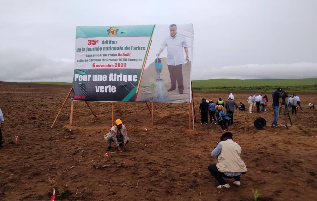 L'image montre un vaste champ de terre nue, où des personnes s'affairent à planter de jeunes arbres. Au centre, un grand panneau affiche le titre "35e édition de la journée nationale de l'arbre" avec une illustration d'un homme en train d'arroser un arbre. La date mentionnée est le 6 novembre 2021, et le slogan "Pour une Afrique verte" est inscrit en bas. Le ciel est nuageux, ce qui suggère un temps frais et peut-être humide, et l'atmosphère semble dynamique, les participants se réunissant pour contribuer à cette initiative de reforestation.