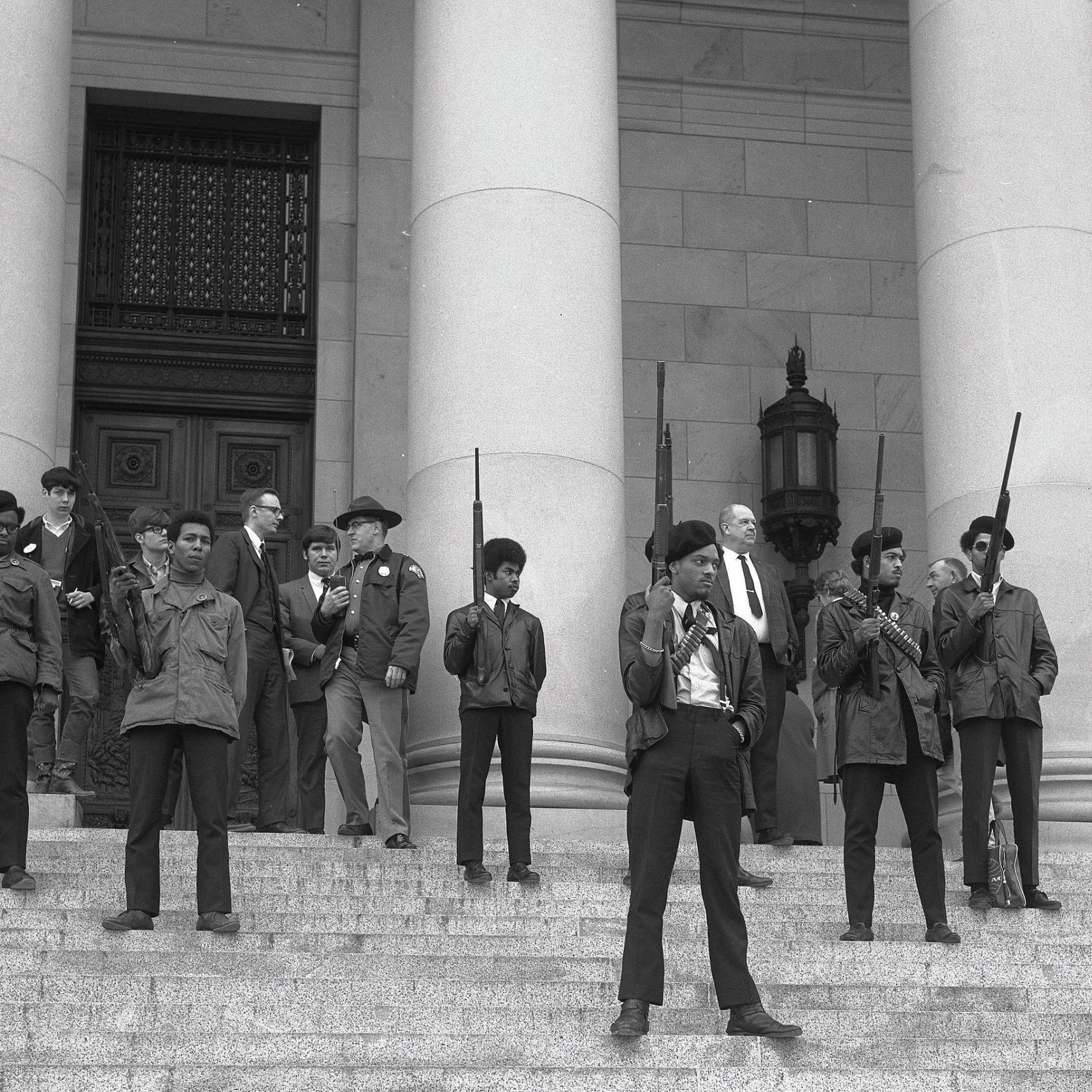 L'image montre un groupe de personnes sur les marches d'un bâtiment imposant, probablement une institution gouvernementale. Au premier plan, plusieurs jeunes hommes se tiennent debout, portant des vestes sombres et des armes à feu, avec des expressions déterminées sur leurs visages. Ils sont entourés d'autres individus, certains vêtus de manière formelle, comme des costumes et des chapeaux. Tous semblent engagés dans une sorte de manifestation ou de rassemblement, émettant un sentiment de tension et de détermination. L'architecture du bâtiment est majestueuse, avec de grandes colonnes en arrière-plan. L'atmosphère générale de l'image évoque une période de lutte pour les droits civiques.