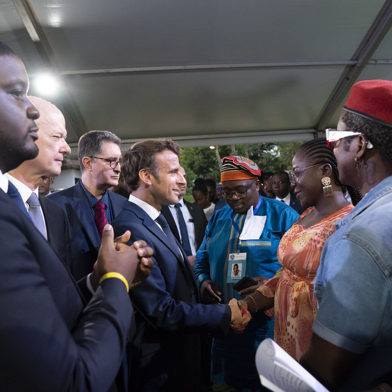 L'image représente un groupe de personnes réunies sous un chapiteau. Au centre, un homme en costume sombre, probablement un leader ou une personnalité importante, est en train de serrer la main d'une femme habillée de façon colorée. On peut apercevoir des visages souriants et une ambiance chaleureuse. D'autres personnes, habillées dans des tenues variées, entourent le couple, regardant avec intérêt. Les arrière-plans sont légèrement flous, mais on ressent une atmosphère de camaraderie et d'échange.