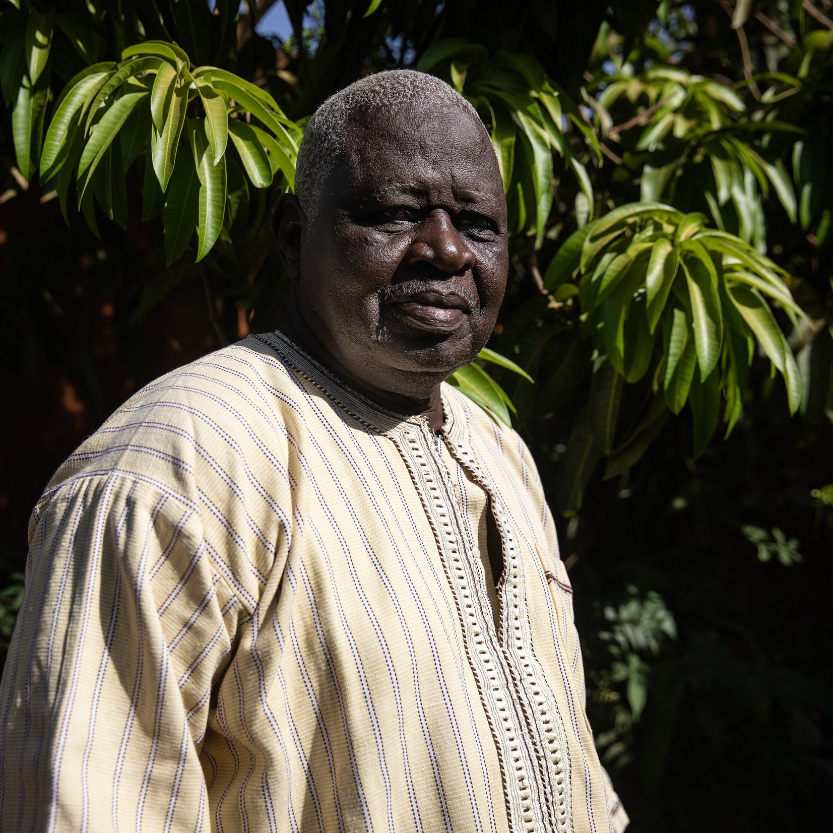 L'image présente un homme debout dans un environnement extérieur verdoyant. Il porte une tunique ample de couleur claire, ornée de motifs discrets, qui témoigne d'un style traditionnel. Son visage est marqué par des traits qui dégagent une certaine sagesse et sérénité. Les cheveux de l'homme sont grisonnants, ajoutant à son apparence mature. En arrière-plan, on aperçoit un feuillage dense et luxuriant, créant un contraste avec sa tenue. La lumière naturelle met en valeur les détails de son visage et de son vêtement, tout en évoquant une atmosphère paisible et accueillante.