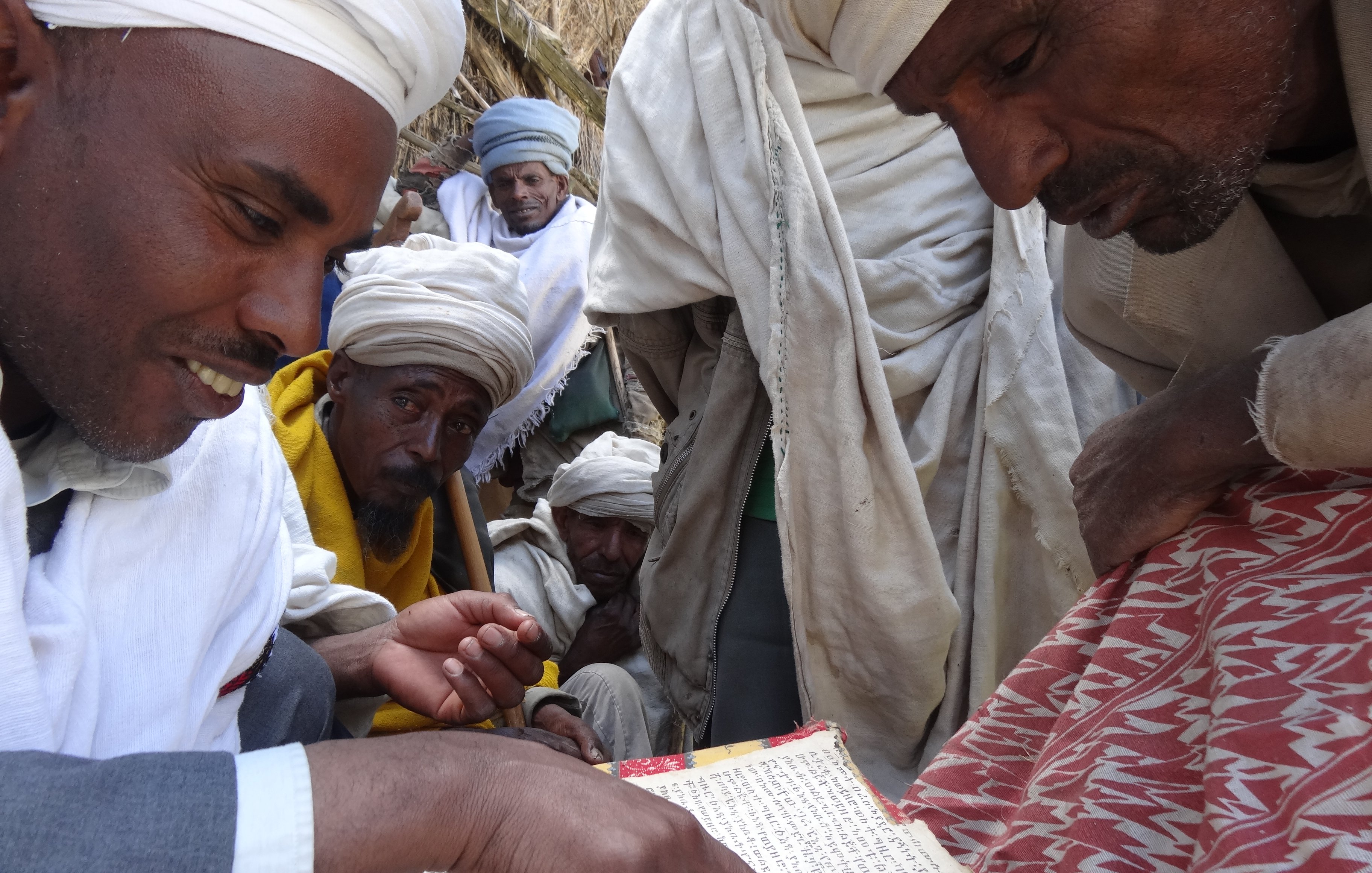 L'image montre un groupe de personnes, principalement des hommes, qui se rassemblent autour d'une table basse sur laquelle se trouve un ancien manuscrit. Un homme au premier plan, portant un habit traditionnel et un turban, montre du doigt une partie du texte. Les autres hommes, qui semblent intéressés, l'observent attentivement. Ils portent également des vêtements traditionnels, et leur expression trahit un mélange de curiosité et de respect. L'ambiance de la scène évoque un moment de partage et de transmission de connaissances, dans un cadre simple mais chargé de culture. En arrière-plan, des éléments naturels et des textures de tissus ajoutent une profondeur à l'atmosphère.