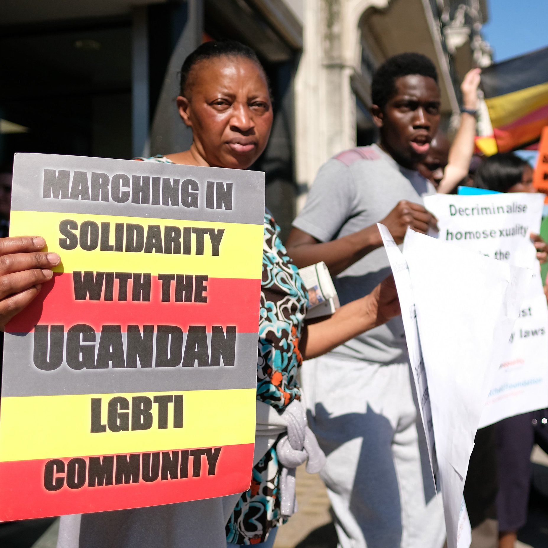 Dans cette image, un groupe de personnes participe à une marche de solidarité en faveur de la communauté LGBTI ougandaise. Les manifestants tiennent des pancartes avec des messages tels que "Marchons en solidarité avec la communauté LGBTI ougandaise" et "Décriminalisez l'homosexualité". L'ambiance est énergique et déterminée, et le groupe affiche une grande diversité. En arrière-plan, on peut voir un drapeau de l'Ouganda flapper, créant un contraste visuel avec les affiches et les vêtements des participants. Les personnes expriment un fort engagement pour les droits humains et l'égalité.