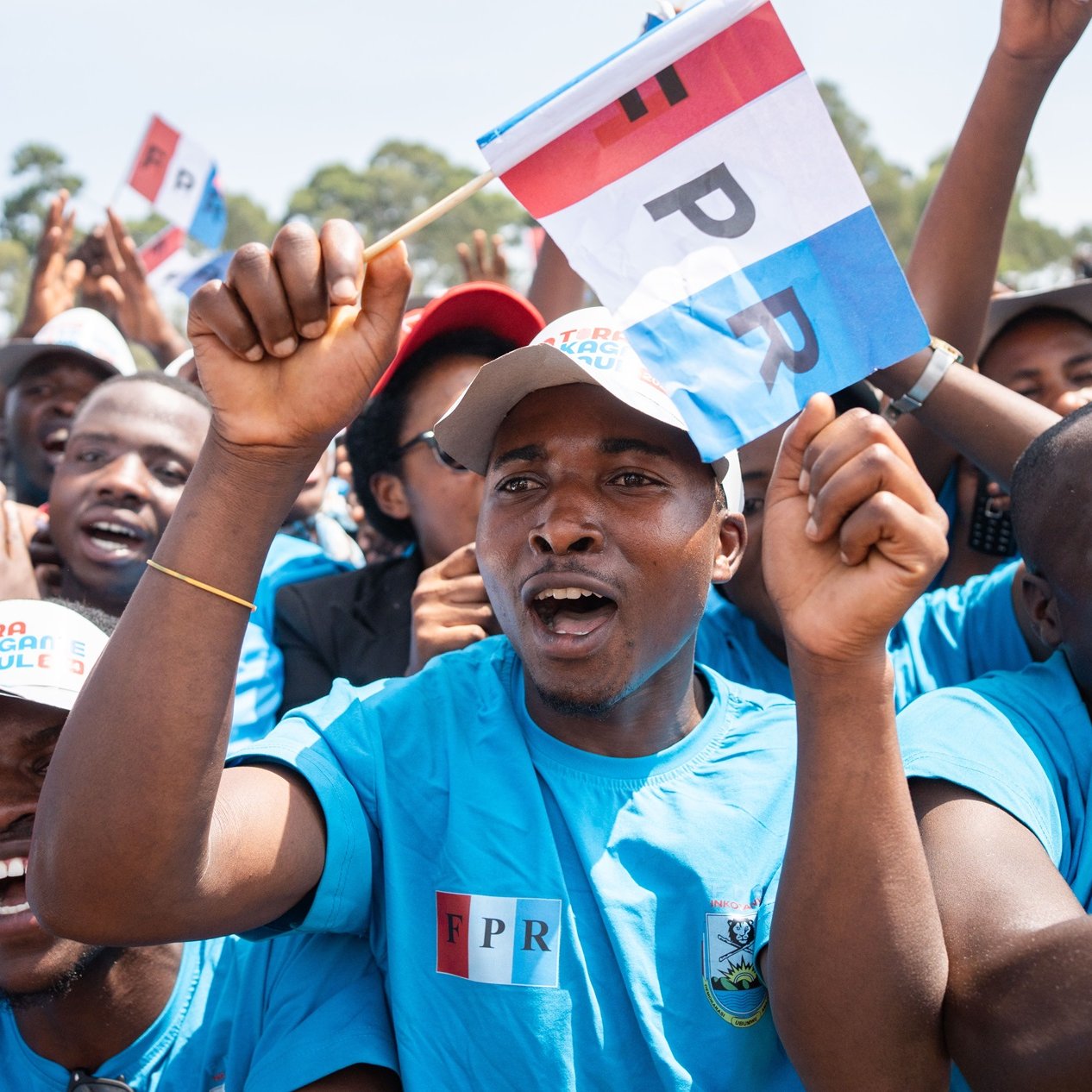 L'image montre un groupe de personnes en train de célébrer ou de soutenir une cause. Les participants portent des t-shirts bleus, et certains brandissent des drapeaux aux couleurs vives, portant l'inscription "FPR". Leur visage exprime de la passion et de l'enthousiasme, avec des sourires et des cris de joie. En arrière-plan, on aperçoit d'autres supporters, créant une atmosphère festive et animée. L'environnement semble ensoleillé, et l'énergie collective est palpable, illustrant un moment de solidarité et de camaraderie.
