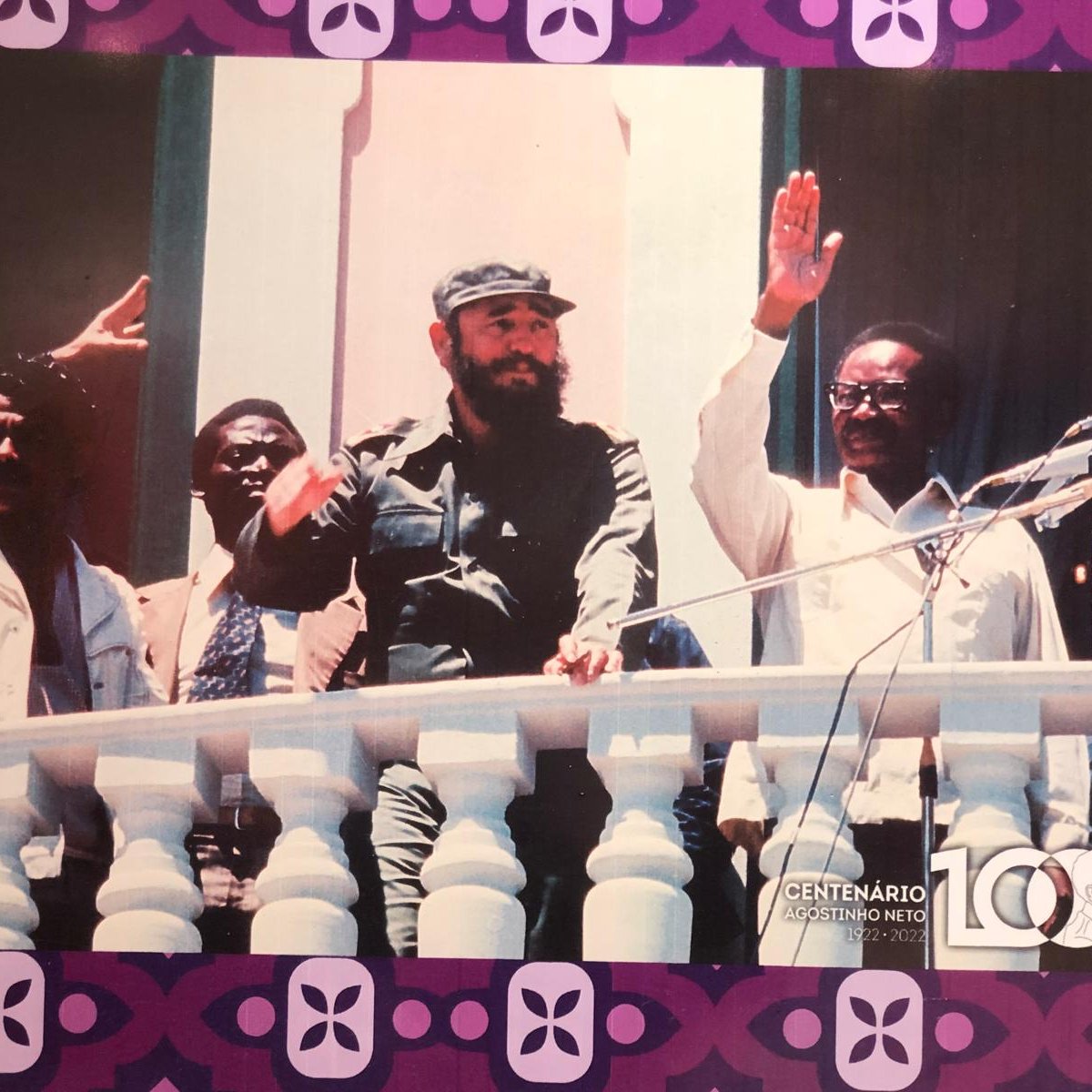 L'image représente un groupe de personnes sur un balcon, probablement lors d'un événement politique. Au centre, un homme avec une barbe, portant un uniforme militaire et un chapeau, lève la main en signe de salutation. À sa gauche, un autre homme en chemise claire semble aussi s'adresser à la foule. À droite, un homme en costume fait un geste similaire. En arrière-plan, on peut voir des murs peints dans des teintes claires, et le décor est encadré par des motifs colorés à la base de l'image. L'atmosphère semble festive et engageante.