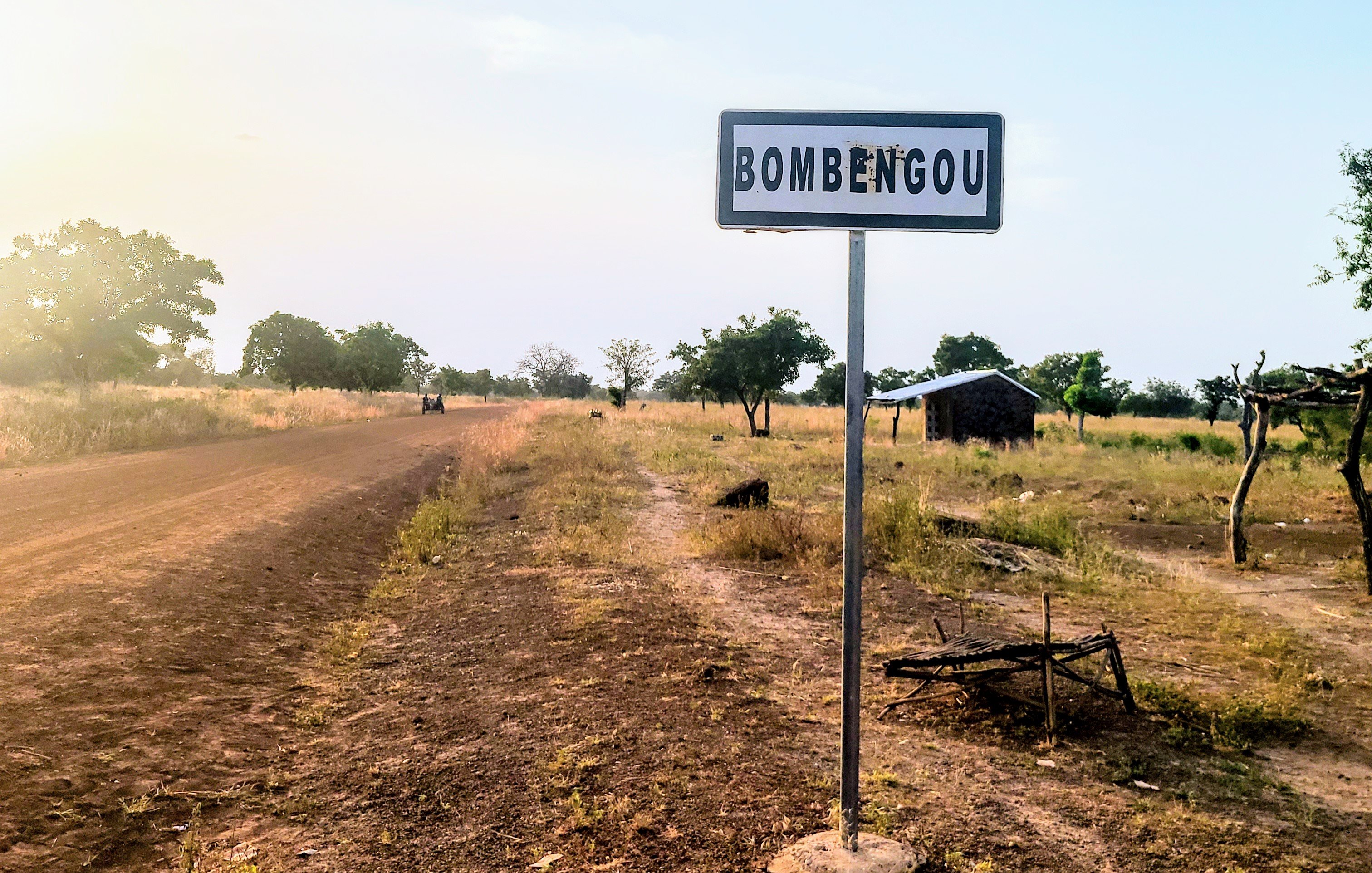 Cette image montre un panneau de signalisation indiquant le nom d'un village, "Bombengou", situé sur un chemin rural. Ce chemin est bordé de terre et de végétation, avec des arbres dispersés le long des côtés. La lumière du soleil crée une atmosphère douce et chaleureuse, suggérant un moment de la journée où la luminosité est agréable. En arrière-plan, on peut apercevoir une petite construction rudimentaire, peut-être une maison ou un abri. L'environnement semble calme et naturel, typique de la campagne.