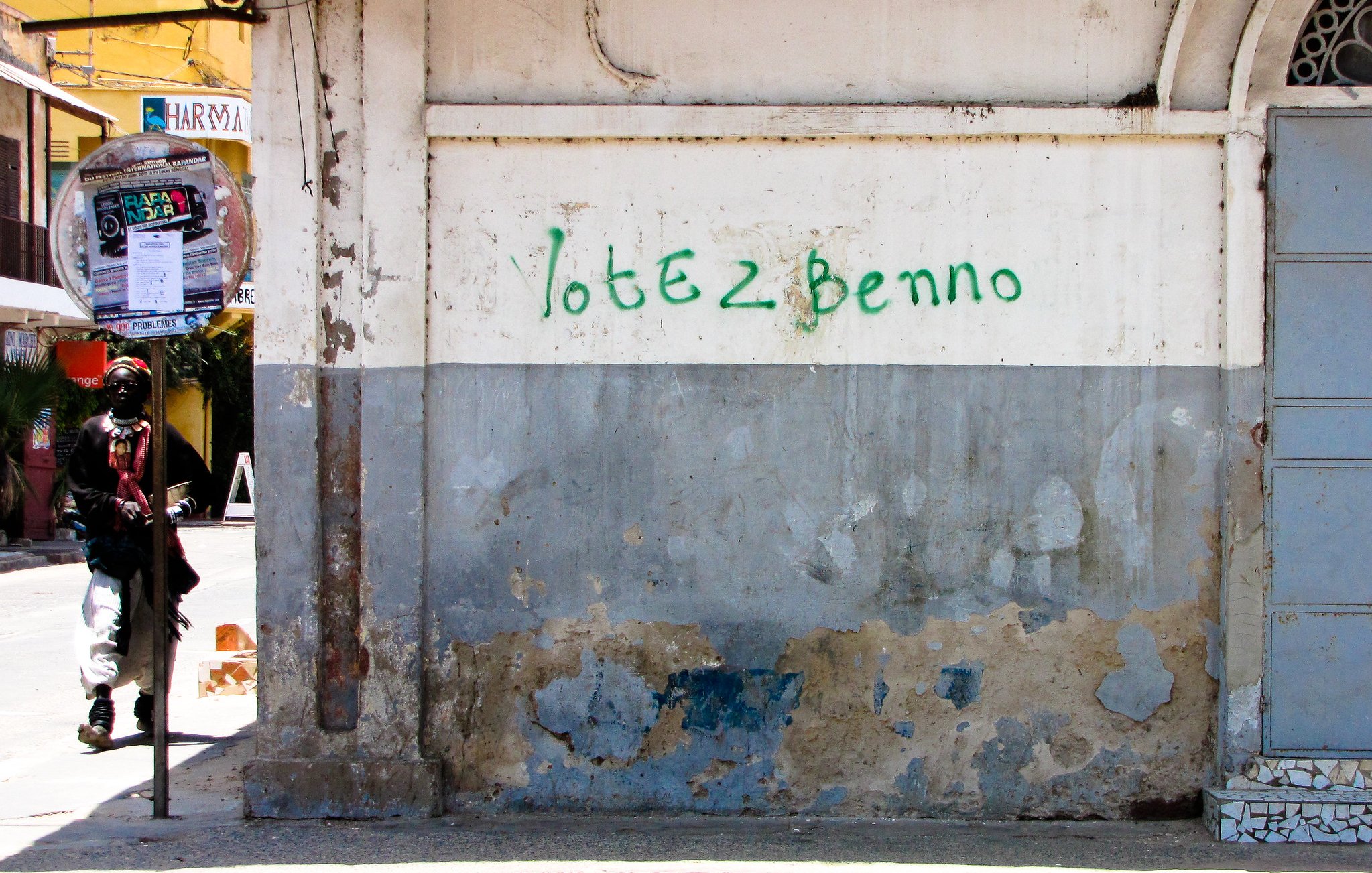 Cette image montre un mur extérieur dans une rue urbaine. La surface du mur est usée, avec de la peinture écaillée et des marques d'usure qui révèlent le temps qui passe. Sur la partie supérieure du mur, il y a une inscription verte qui dit "Votez Benno". À côté du mur, un homme est visible de dos, appuyé contre le mur. Son style vestimentaire semble décontracté. En arrière-plan, on peut apercevoir un peu de végétation et d'autres bâtiments qui enrichissent le paysage urbain. L'atmosphère générale évoque une scène de la vie quotidienne dans une ville animée.