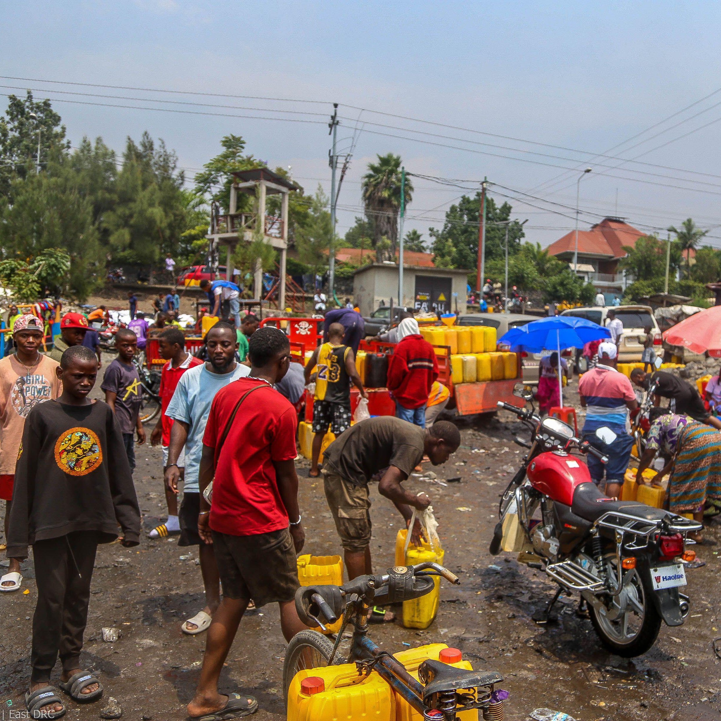 The image depicts a crowded scene in an outdoor setting, likely a market or gathering place. People are seen collecting water from yellow jerry cans, some of which are stacked nearby. The atmosphere appears busy and somewhat chaotic, with individuals interacting and engaging in various activities. There are motorcycles and umbrellas visible, suggesting a warm, sunny environment. The ground is littered with debris, indicating a lack of cleanliness in the area. The background features buildings and trees, contributing to the urban landscape.