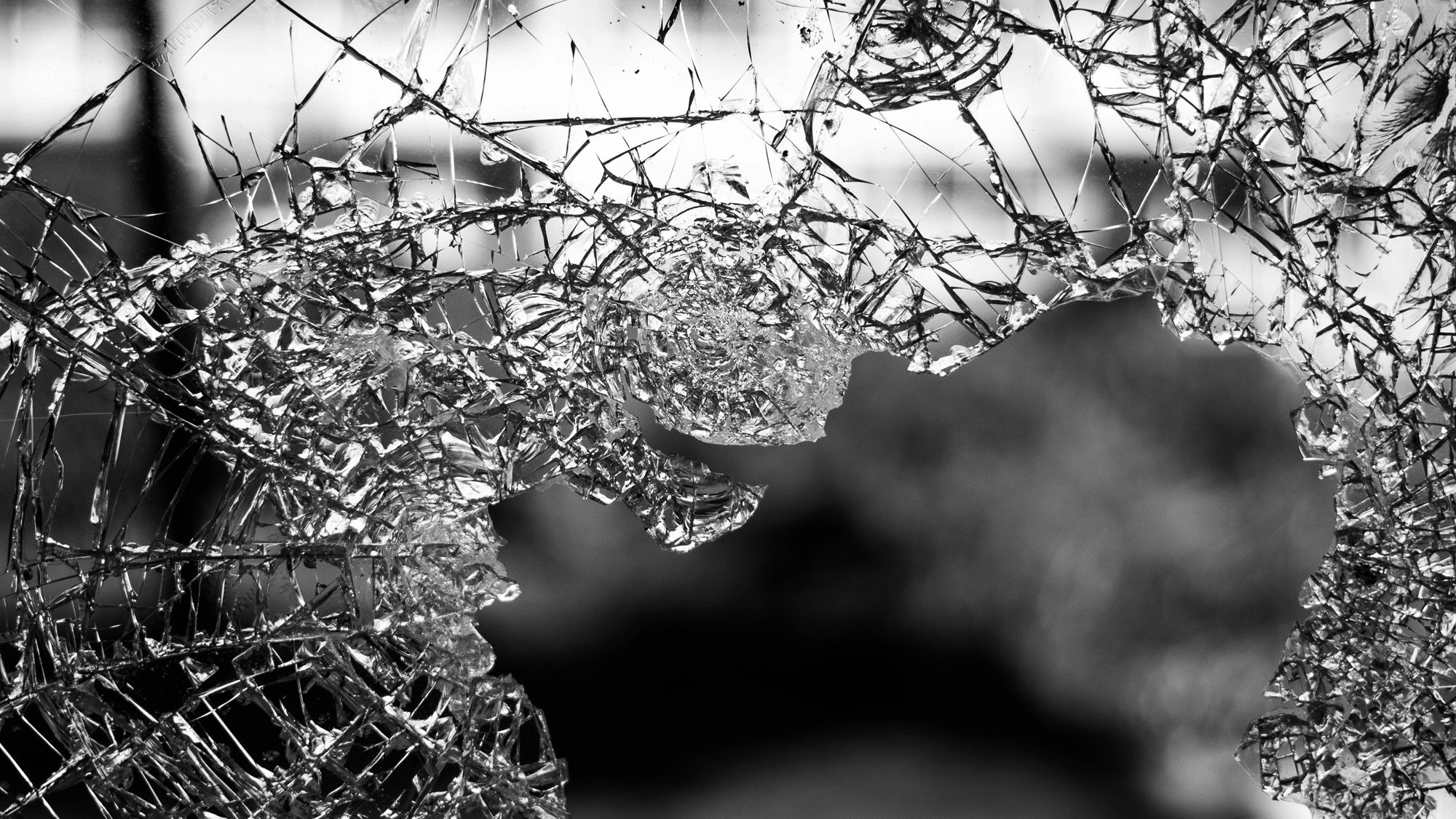L'image montre un morceau de verre brisé, dont les nombreux éclats forment des motifs irréguliers. On peut ressentir la fragilité et la netteté des bords du verre. La lumière semble traverser certains éclats, créant des jeux d'ombre et de lumière. L'arrière-plan est flou, évoquant une ambiance sombre et mystérieuse. Cette composition souligne la beauté inattendue du verre cassé, tout en évoquant une sensation de vulnérabilité.