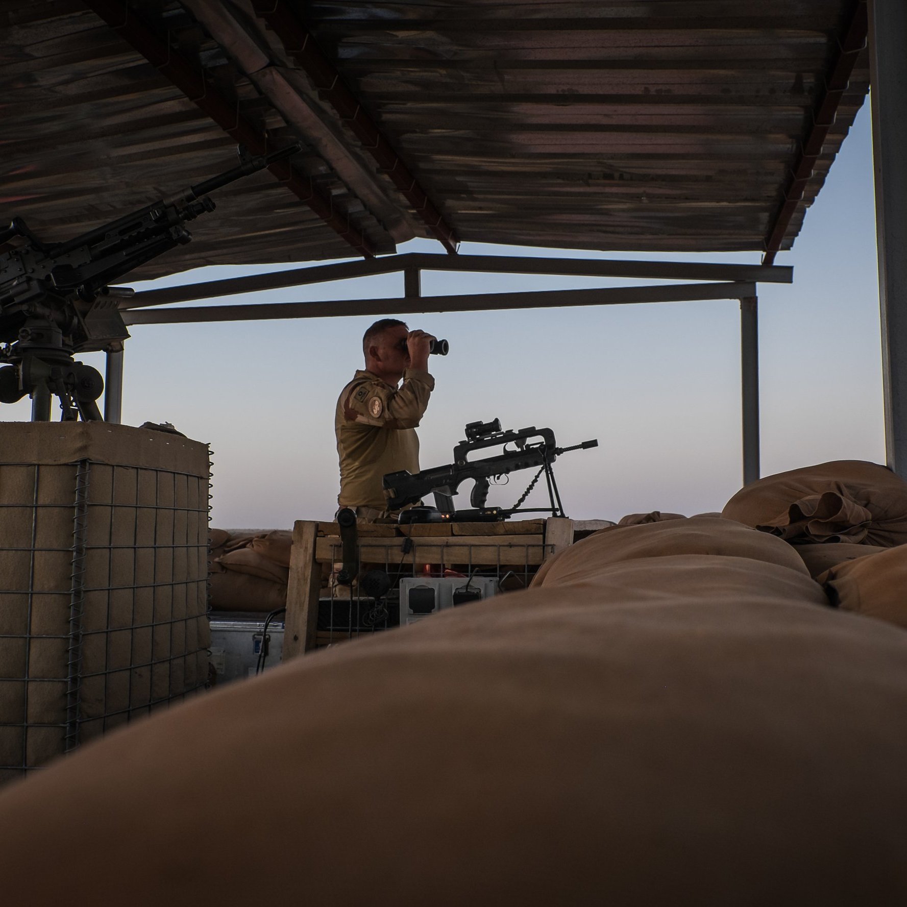 L'image montre un soldat en position d'observation sur un poste de garde en hauteur. Il se tient debout, avec des jumelles à la main, scrutant l'horizon. À sa droite, on aperçoit une mitrailleuse montée sur un support. Le sol est recouvert de sacs de sable, utilisés comme protection. L'éclairage de la scène évoque un moment de la journée où le ciel est teinté de couleurs douces, créant une atmosphère calme malgré le contexte militaire. Le soldat porte un uniforme et semble attentif à son environnement.