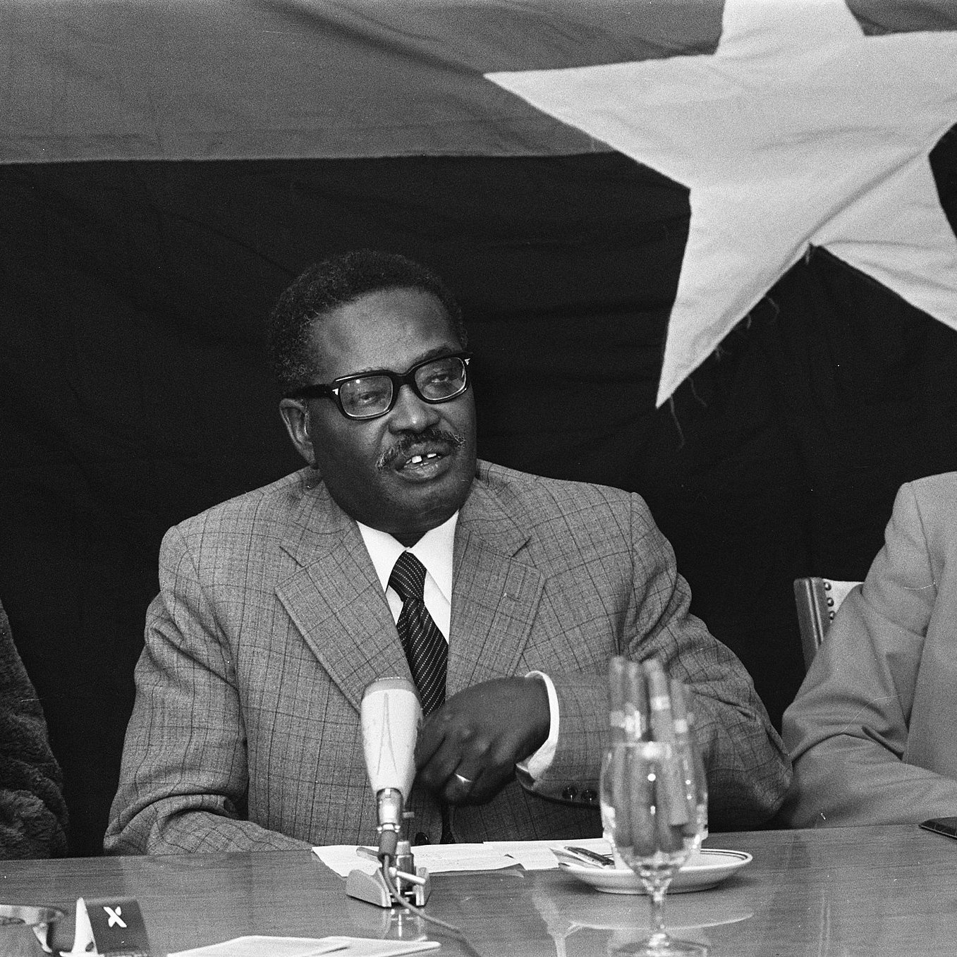 L'image montre une réunion où deux hommes sont assis à une table. À gauche, un homme d'âge moyen, portant des lunettes et un costume gris à carreaux, semble s'adresser aux autres participants, gesticulant légèrement avec une main. Il a une expression attentive et engagée. À sa droite, un homme plus âgé avec des cheveux gris et des lunettes, habillé d'un pull noir et d'une veste beige, l'écoute avec intérêt. En arrière-plan, on devine un grand drapeau noir avec une étoile blanche, ajoutant un contexte politique à la scène. Sur la table, il y a quelques verres, un microphone et un bloc-notes. L'atmosphère semble sérieuse et concentrée.