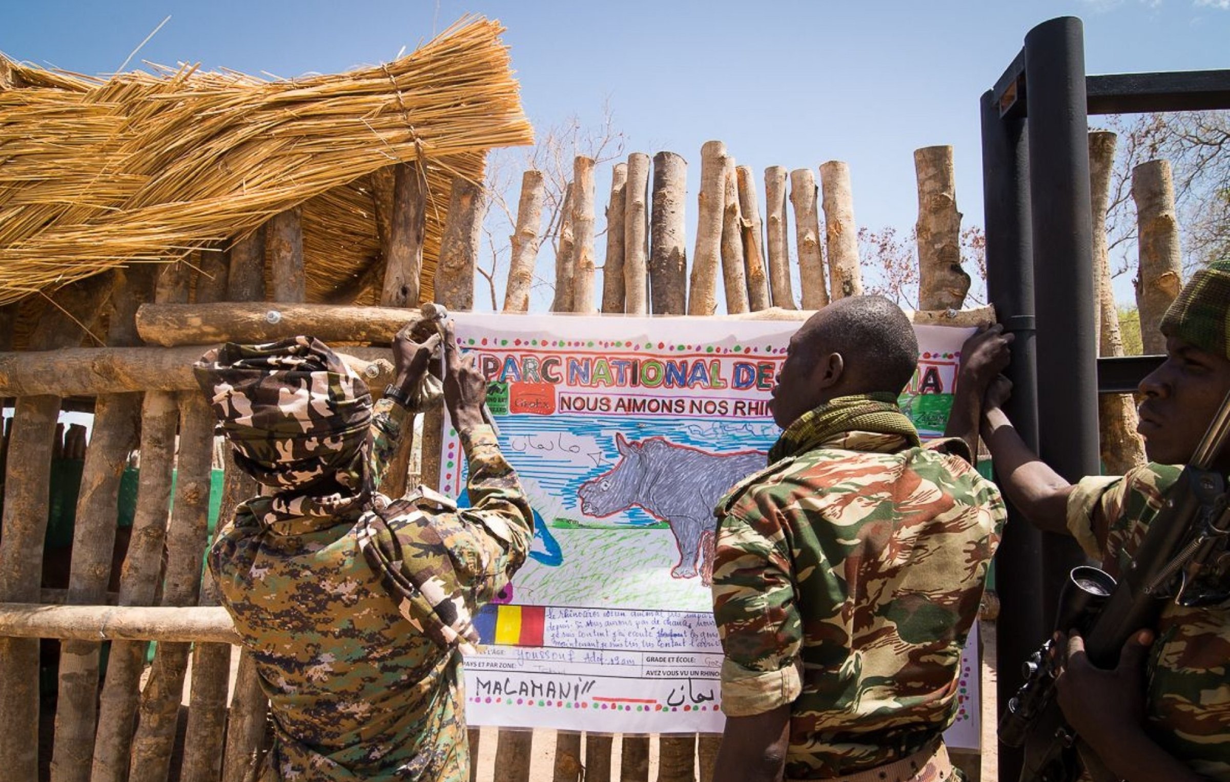 Sur l'image, nous voyons deux soldats en uniforme coloré, engagés dans une action collective. Ils sont en train d'accrocher une grande affiche sur un mur en bois fait de branches. L'affiche représente un rhinocéros, avec un dessin coloré et un message écrit en français qui exprime un engagement envers la protection de cette espèce. Le cadre est en plein air, avec un ciel bleu et quelques éléments naturels visibles. L'atmosphère dégage un sentiment de protection et d'éducation environnementale, soulignant l'importance de préserver la faune.