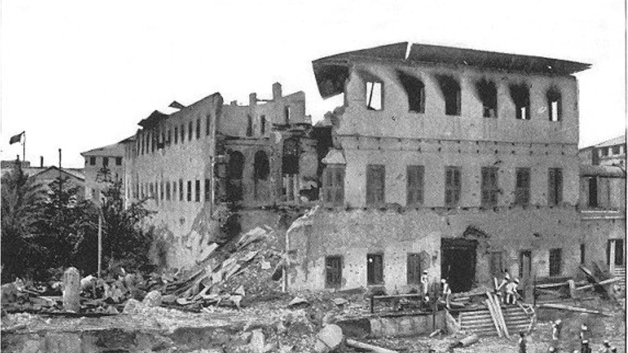 L'image montre un bâtiment sévèrement endommagé, probablement à la suite d'un tremblement de terre ou d'une catastrophe. La façade du bâtiment est partiellement effondrée, révélant des murs fissurés et des fenêtres brisées. Des débris sont éparpillés autour, créant un paysage chaotique. Au premier plan, on peut voir un groupe de personnes qui semblent examiner les dégâts, échangeant probablement leurs impressions. L'environnement est empreint d'une atmosphère de désolation, illustrant les conséquences d'un événement tragique sur l'architecture et la communauté environnante.