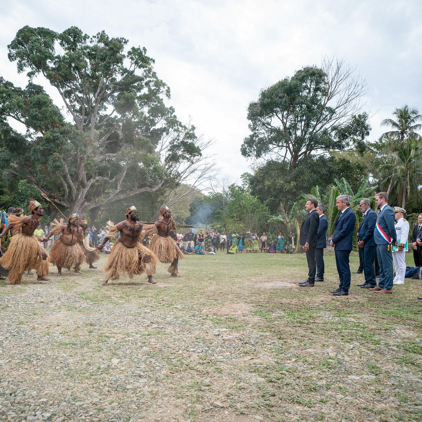 L'image montre un groupe de personnes en tenue traditionnelle, qui semblent participer à une cérémonie ou une célébration. Au centre, des hommes vêtus de costumes en fibres naturelles dansent avec énergie. Ils sont entourés par un public, composé de personnes en tenue formelle, qui observe attentivement la scène. L'environnement est verdoyant, avec des arbres et une légère brume qui donne une atmosphère presque mystique. À l'arrière-plan, un groupe de spectateurs semble apprécier l'événement, ajoutant une dimension communautaire à cette célébration culturelle.