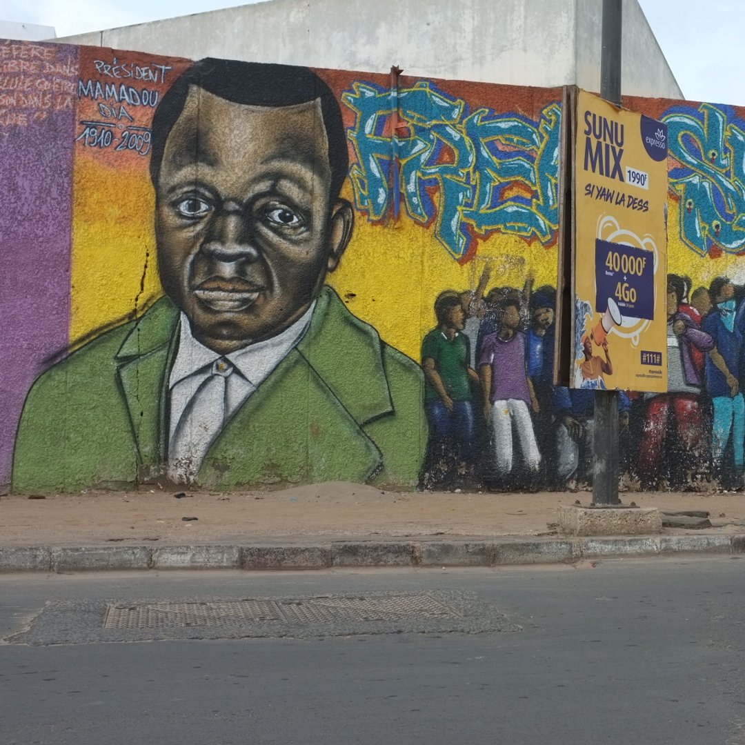 L'image montre un mur de graffitis vibrant et coloré. Au centre, il y a un portrait d'une personne au visage sérieux, avec des traits marqués, habillée d'un costume vert. Autour de ce portrait, différentes figures sont représentées, suggérant une scène animée de personnes rassemblées, peut-être en train de discuter ou de célébrer. À côté, on peut voir une affiche collée sur le mur avec des éléments graphiques. En bas de l'image, un taxi à la carrosserie orange et noir passe sur la route, ajoutant une touche de dynamisme à la scène urbaine. L'ensemble évoque une ambiance de vie communautaire et de culture urbaine.