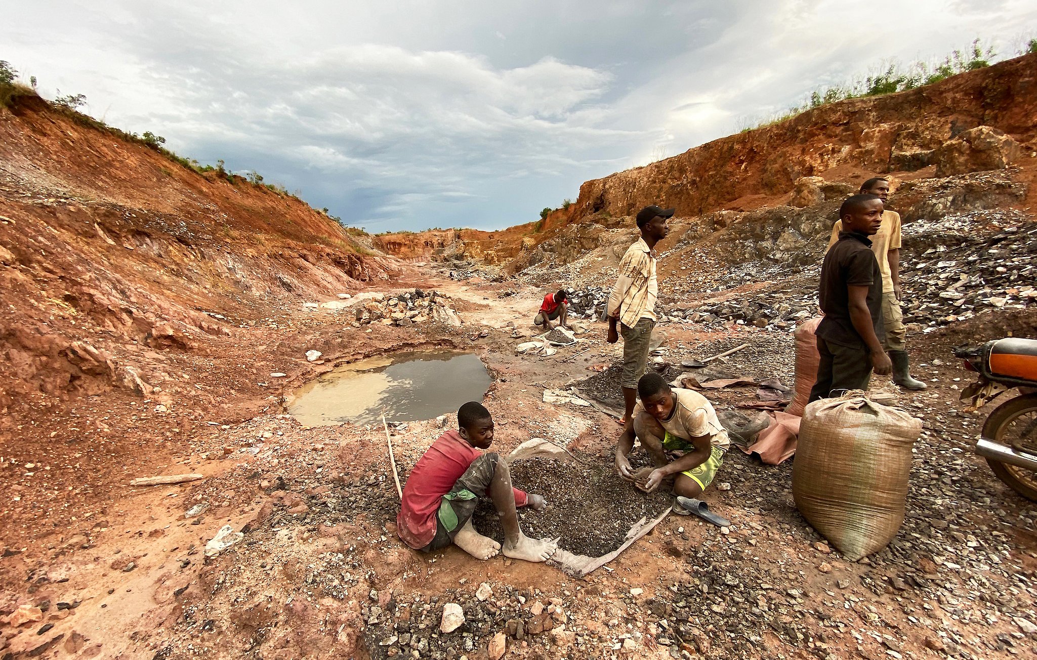 L'image montre une scène rurale d'exploitation minière. On y voit un paysage marqué par des fouilles, avec un sol rouillé et des montagnes de terre séparées. Au premier plan, plusieurs personnes, principalement des hommes et des jeunes garçons, sont en train de travailler. Certains sont accroupis, tamisant des pierres ou ramassant des minéraux, tandis que d'autres semblent transporter des sacs pleins de matériaux. À gauche, il y a un petit plan d'eau, qui est trouble, entouré de débris miniers. Le ciel est nuageux, ajoutant une ambiance sombre à la scène, évoquant un travail difficile et épuisant.