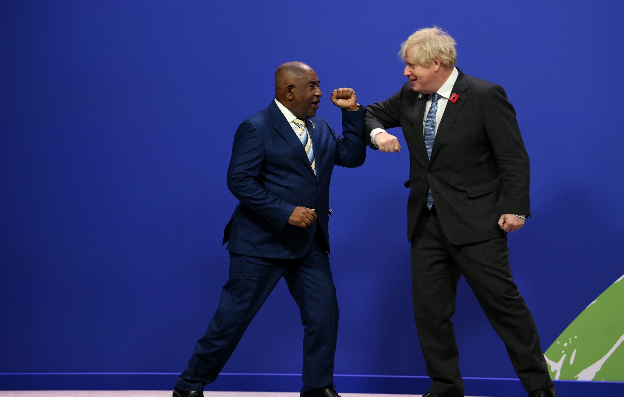 Dans cette image, deux hommes se tiennent sur une scène avec un fond bleu. L'un porte un costume sombre avec une cravate claire, se tenant sur la gauche, et semble sourire en regardant l'autre homme. Ce dernier, sur la droite, est habillé d'un costume noir avec une cravate à motifs et semble également souriant. Les deux hommes portent leurs bras en avant dans un geste de salut amical, touchant légèrement leurs coudes. L'ambiance générale de l'image est dynamique et conviviale, suggérant une interaction positive entre eux.