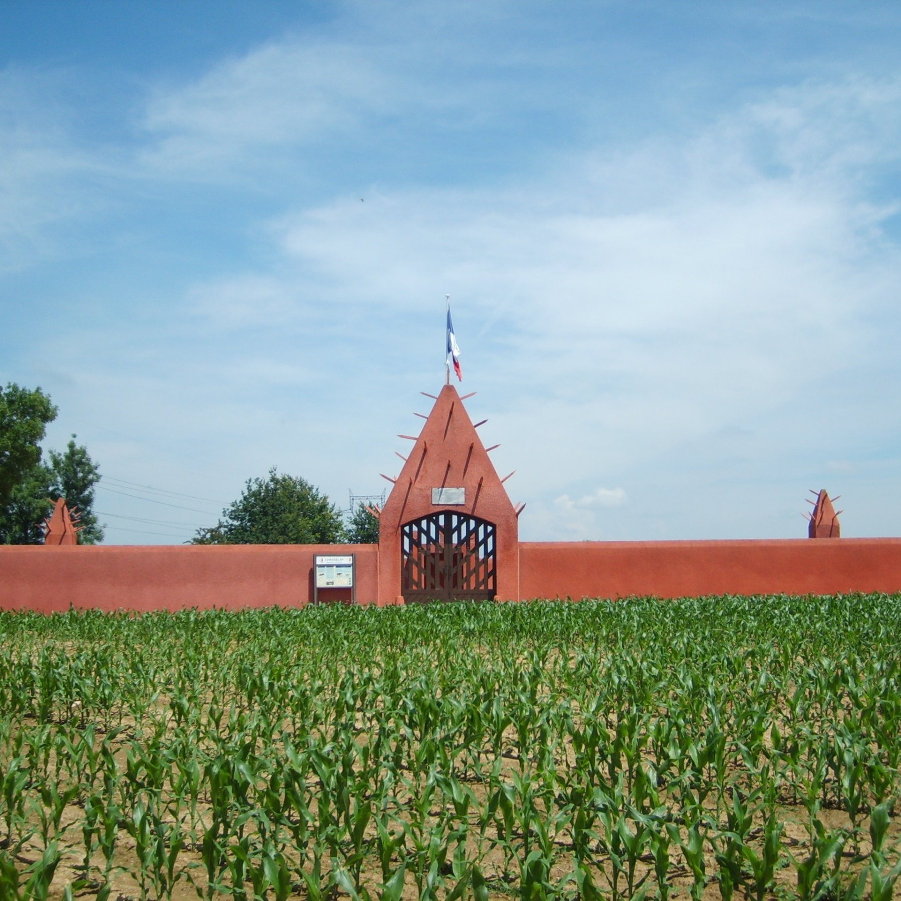 L'image présente une grande entrée de style architectural distinctif, peinte en rouge, qui s'élève au-dessus d'un champ de maïs. La structure est dotée d'un mur haut, orné de pointes qui se projettent vers le ciel, créant un aspect à la fois imposant et décoratif. Au centre, une porte en fer forgé permet d'entrer, et on peut apercevoir un drapeau français flottant au-dessus. Le ciel d'un bleu éclatant, parsemé de nuages légers, contraste avec la verdure des jeunes plants de maïs qui s'étendent devant le mur, ajoutant une touche de fraîcheur à la scène.