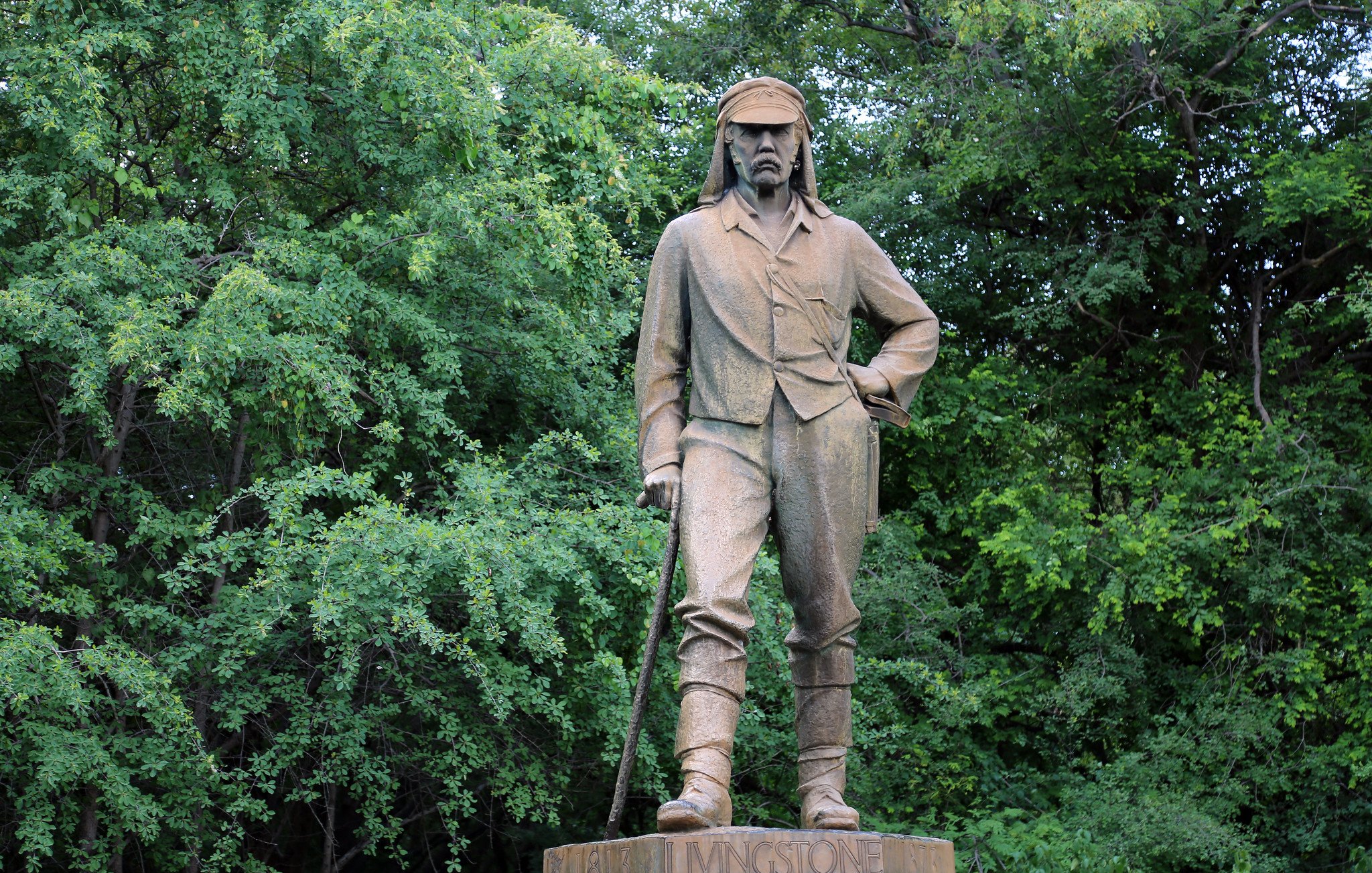 L'image montre une statue en bronze d'un homme qui se tient debout, avec une posture confiante. Il porte des vêtements de style explorateur du XIXe siècle, incluant une chemise à manches longues et un pantalon. Son regard est déterminé et il a une canne à la main, suggérant qu'il pourrait être un aventurier ou un explorateur. La statue est placée sur un socle avec une inscription, et elle est entourée d'une végétation dense, avec des arbres verts en arrière-plan, créant un contraste entre la statue et la nature environnante. L'atmosphère est calme et sereine, reflétant peut-être le respect pour cette figure historique.