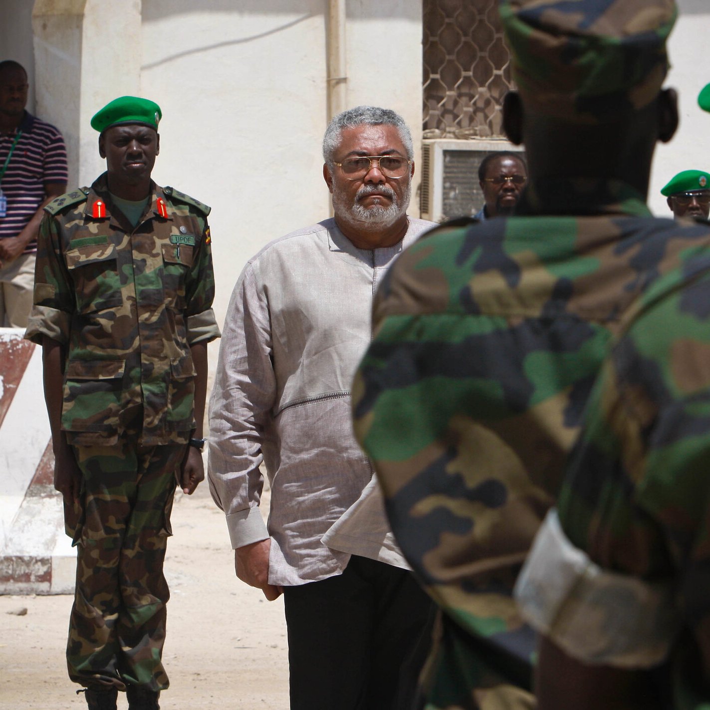L'image montre un homme d'âge moyen au centre, avec des cheveux poivre et sel et une barbe grise. Il porte une chemise claire. Il semble sérieux et concentré, entouré de militaires en uniforme camouflage. Ces soldats, qui se tiennent en rangs, regardent vers lui. En arrière-plan, on peut voir un bâtiment aux murs clairs et un voisinage qui évoque un environnement militaire. L'atmosphère est formelle, soulignant l'importance de la scène.