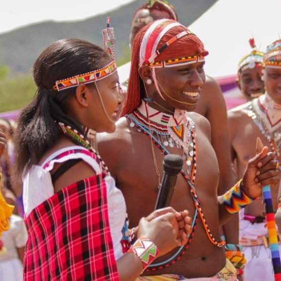 Dans cette image, un groupe de personnes participe à une cérémonie culturelle vibrante. Au premier plan, une femme, vêtue d'une robe traditionnelle colorée avec des motifs, tient un micro et s'adresse à un homme qui se tient à côté d'elle. Cet homme porte un pagne traditionnel et est orné de nombreux accessoires en perles. Ils sont tous deux entourés de plusieurs autres hommes, également en tenue traditionnelle, qui sourient et semblent s'engager dans une conversation joyeuse. Les vêtements sont décorés de motifs colorés et de bijoux en perles, ce qui évoque une atmosphère festive. L'arrière-plan montre des collines verdoyantes, et l'ambiance générale dégage un sens de communauté et de célébration de la culture. L'image respire l'énergie et la convivialité, illustrant des rituels ou des danses traditionnelles.
