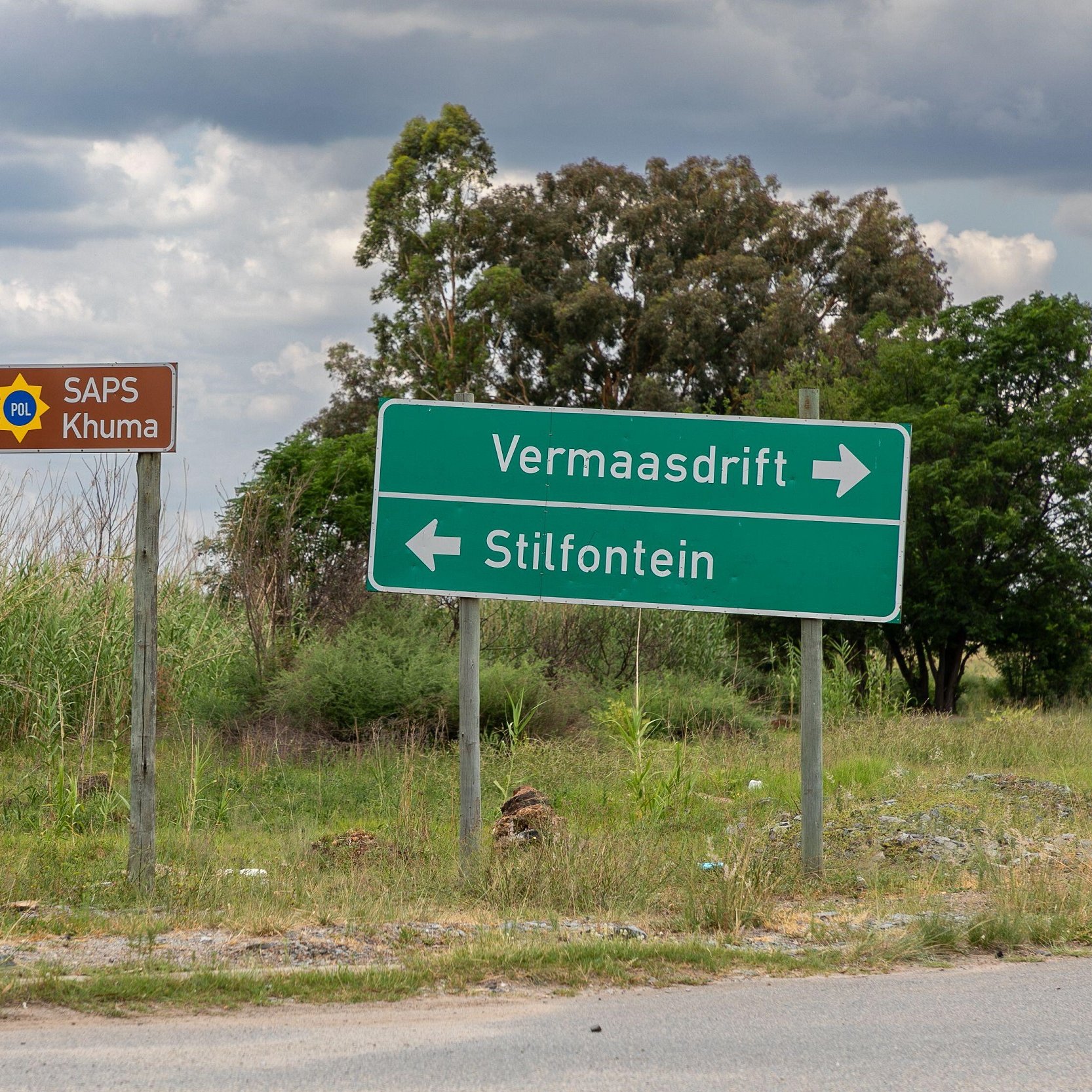 Cette image montre un carrefour routier en extérieur. À gauche, un panneau marron avec un symbole de police indique "SAPS Khuma", suggérant la présence d'un poste de police local. À droite, un panneau vert montre deux flèches directionnelles : une vers "Vermaasdrift" et l'autre vers "Stilfontein". L'environnement est caractérisé par une végétation verte et dense, avec quelques arbres en arrière-plan et un ciel nuageux, créant une atmosphère calme et naturelle. Le sol est asphalté, donnant l'impression d'une route peu fréquentée.